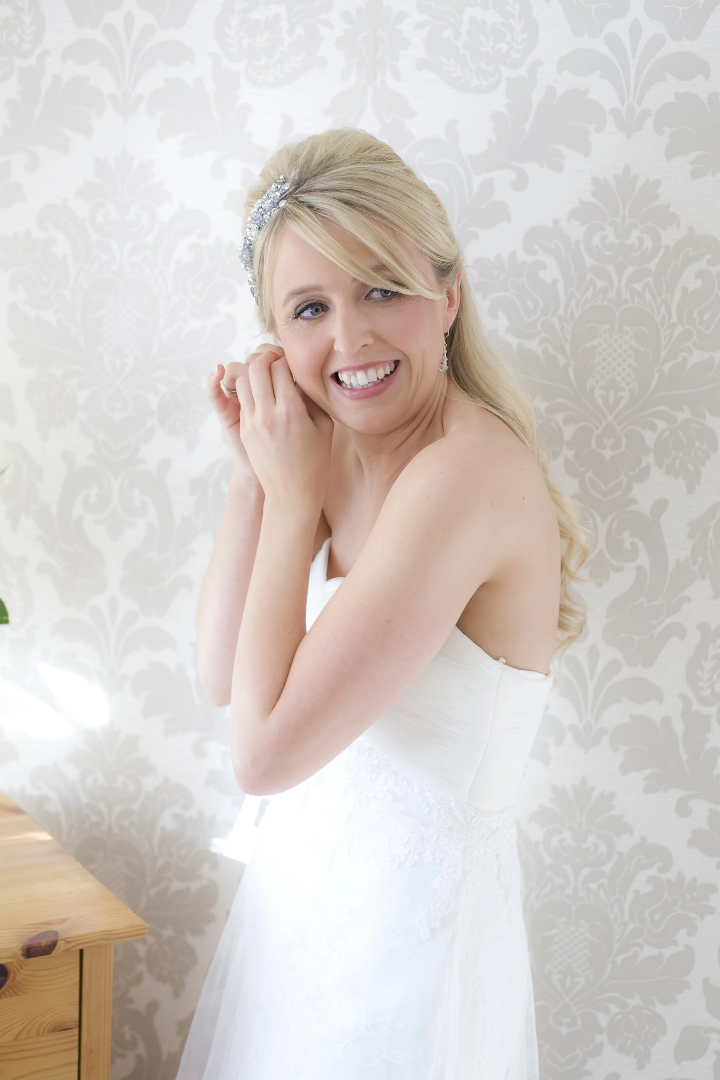 A bride with blonde hair in a strapless white wedding dress, smiling and adjusting her earring, standing in front of a patterned wallpapered wall.