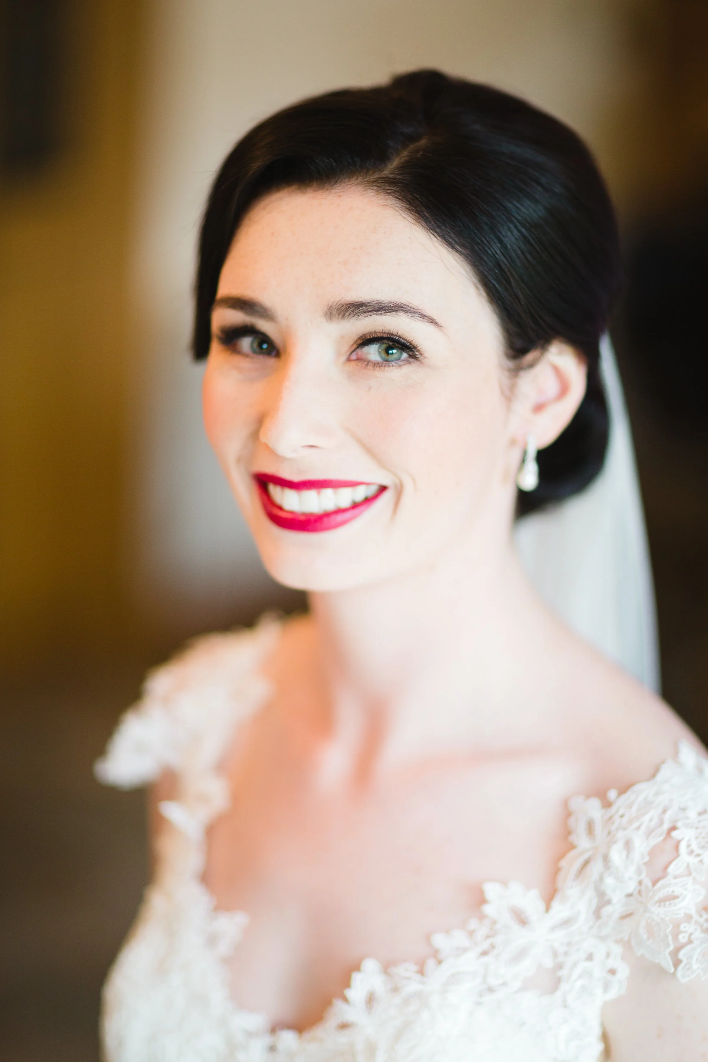 A smiling bride with dark hair and blue eyes, wearing a lace wedding dress and pearl earrings.
