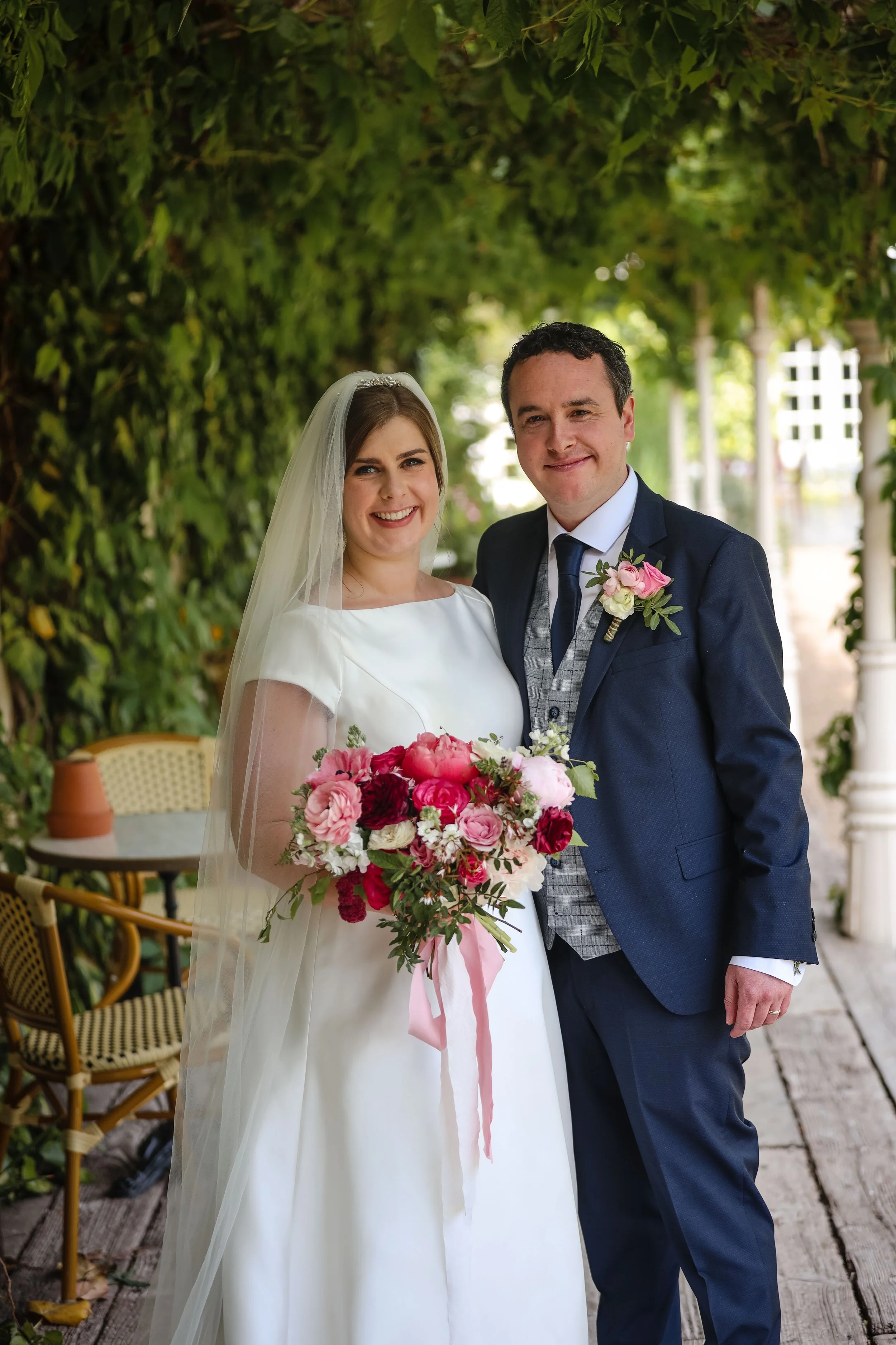 A bride and groom posing together outdoors during their wedding. The bride is holding a bouquet of pink, red, and white flowers. The groom is wearing a navy blue suit with a gray vest and tie, and has a boutonniere matching the bouquet. They are smil