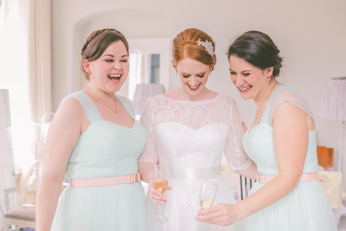 Bride in a white lace wedding dress with friends in light blue dresses celebrating and holding glasses of wine in a bright room.