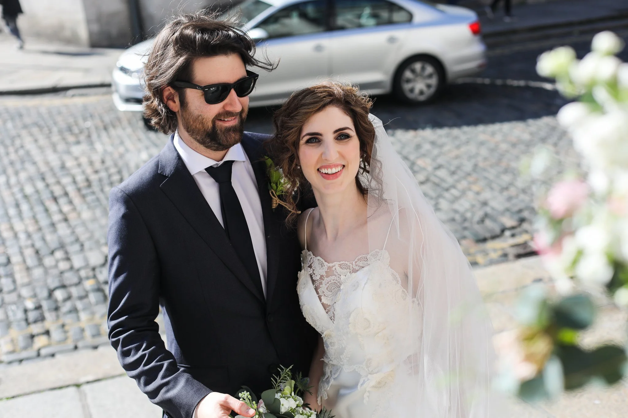 A happy bride and groom smiling for a photo outdoors on a cobblestone street, with cars in the background. The bride is wearing a lace wedding dress and veil, holding a bouquet of white flowers and greenery. The groom is wearing a dark suit with a wh