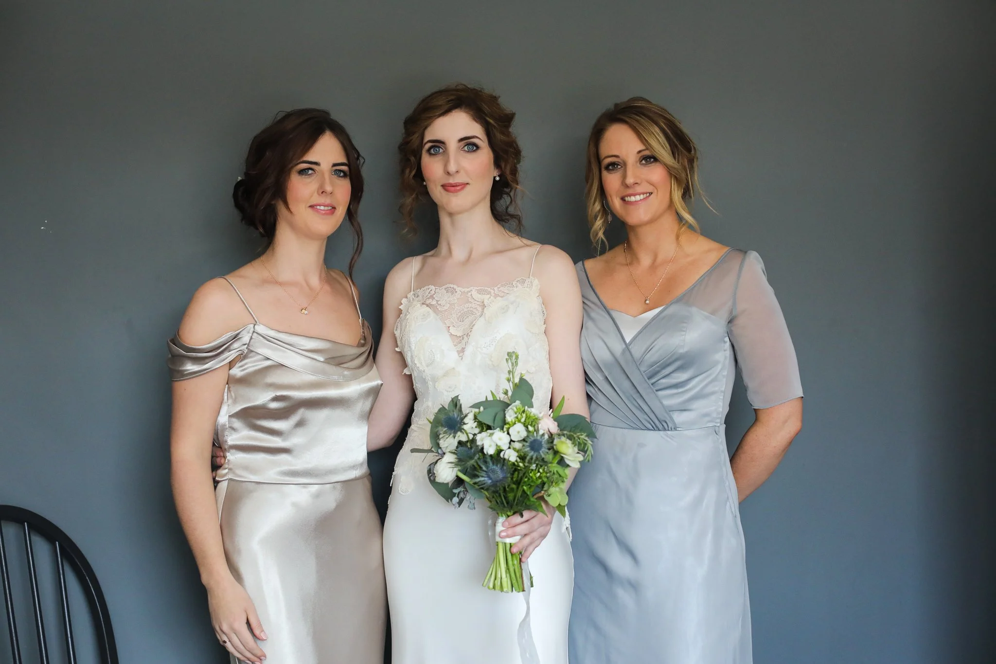 Three women dressed in elegant gowns standing together against a gray wall, with the woman in the middle holding a bouquet of flowers.
