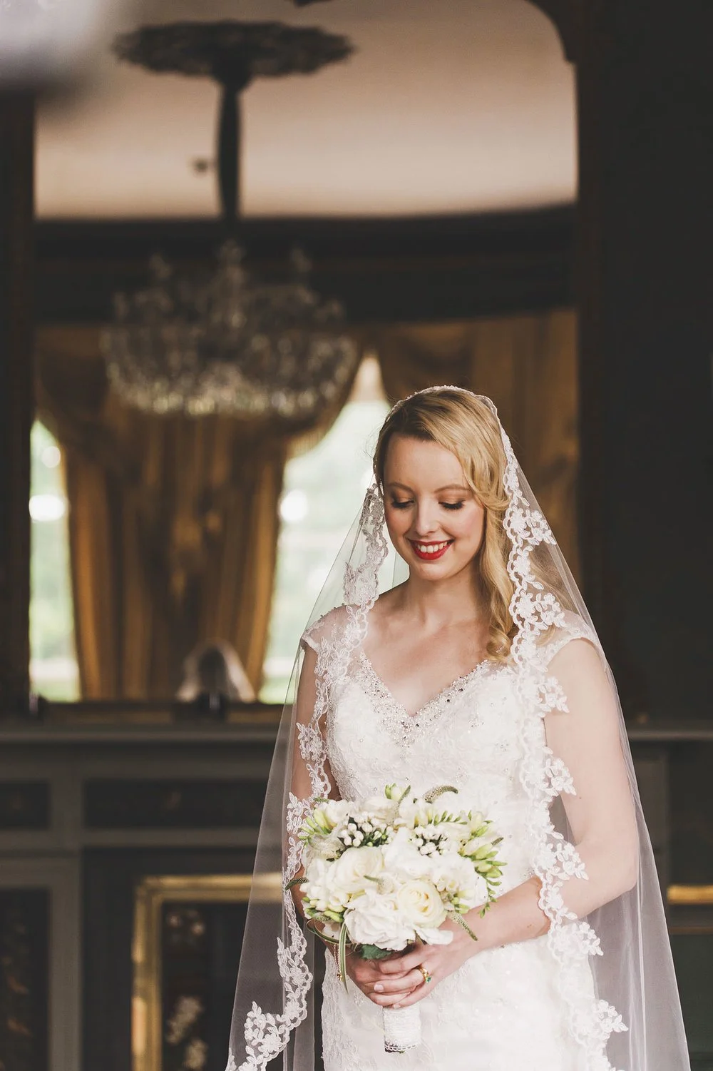 A bride in a lace wedding gown holding a bouquet of white flowers, smiling softly in a luxurious, warmly lit room with large windows and ornate drapes.