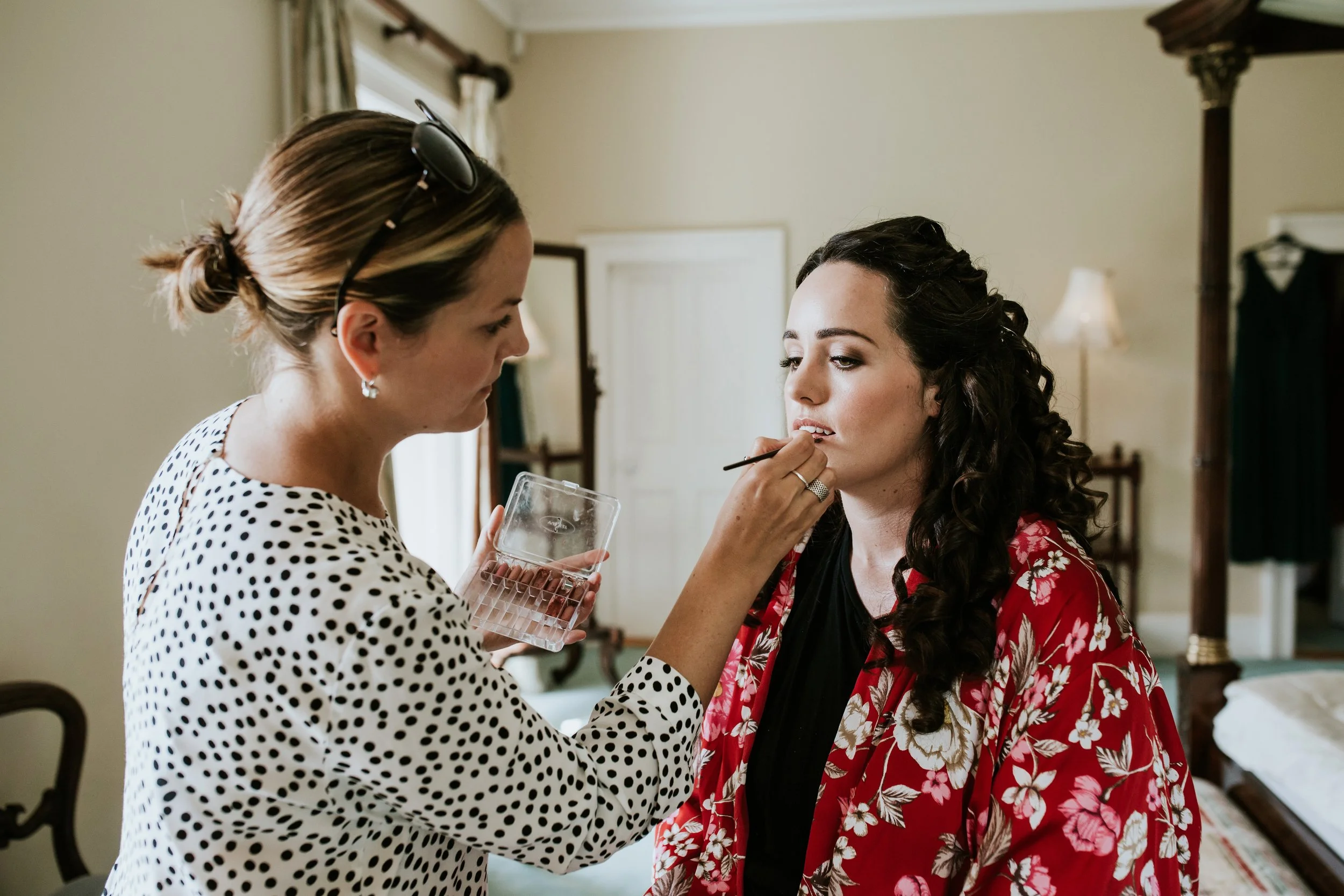 A makeup artist applying lipstick to a woman with long curly hair, wearing a red floral robe, in a bedroom setting.