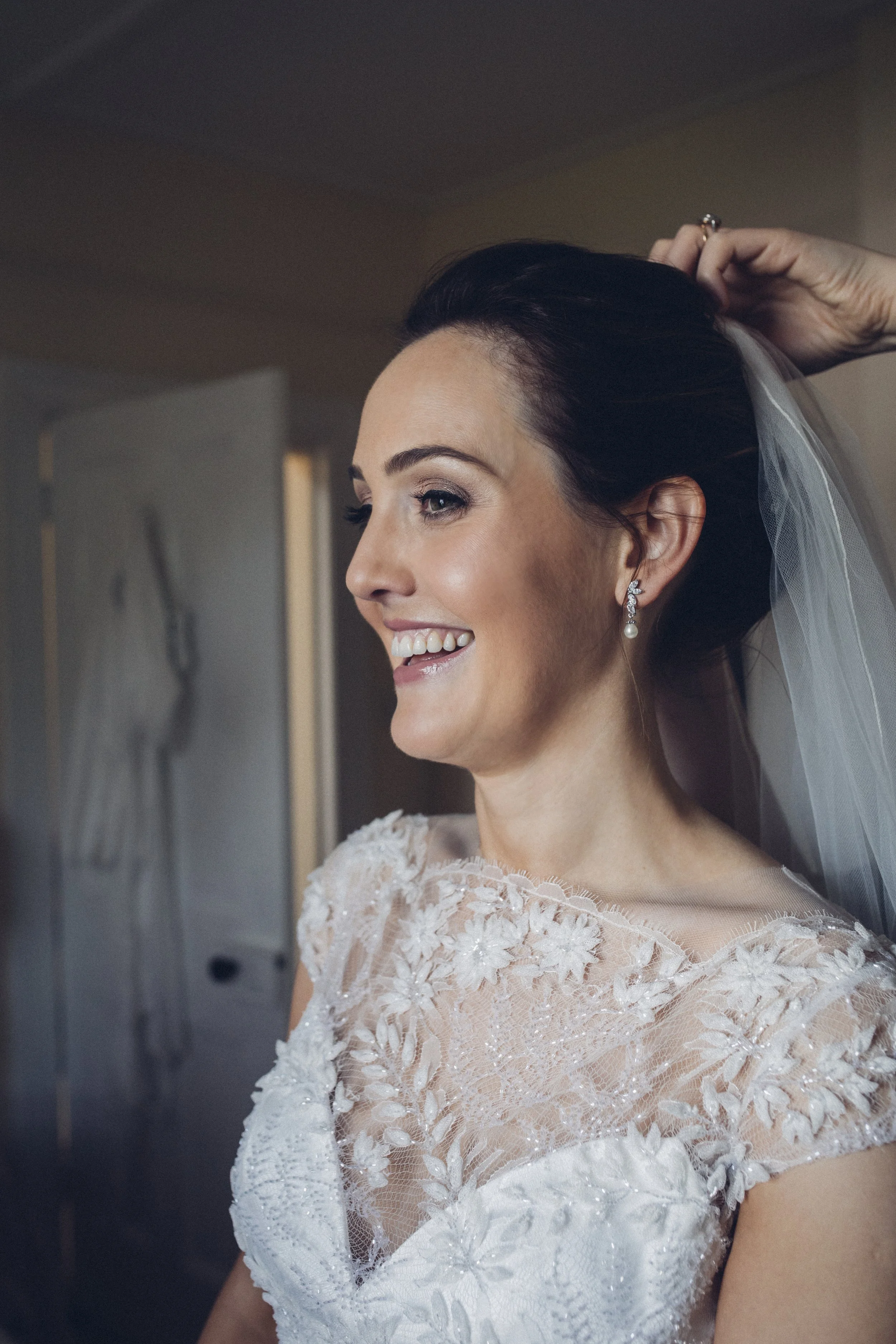 A bride in a wedding dress with lace and floral embroidery, smiling as she adjusts her hair with an accessory, wearing earrings and a veil.