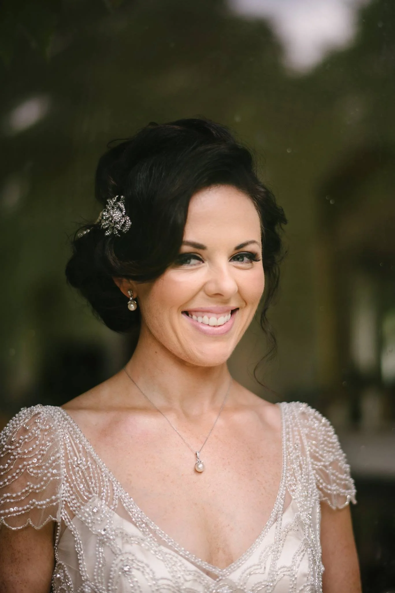 Smiling woman with dark hair styled in an updo, wearing pearl and crystal jewelry and a pearl-embellished dress, standing outdoors.