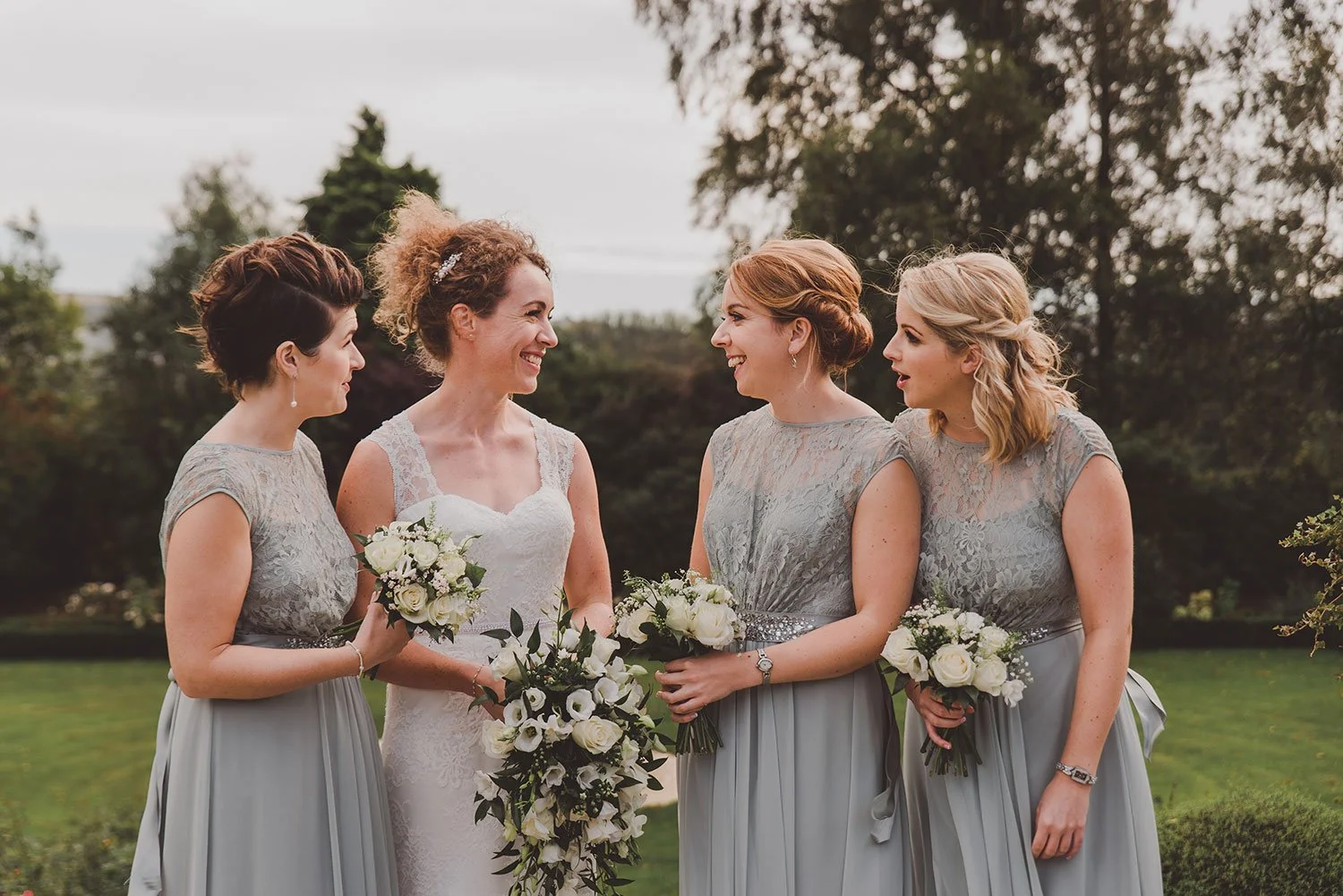 A bride and three bridesmaids standing outdoors, smiling and holding bouquets of white flowers, with trees and a cloudy sky in the background.