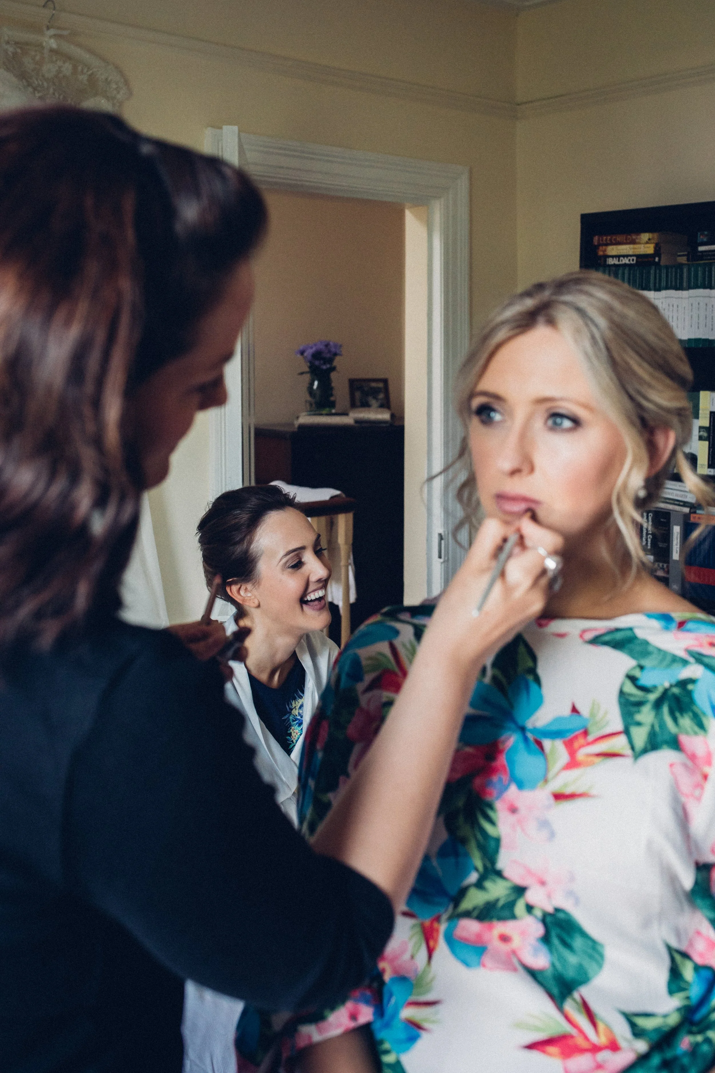 Three women preparing for a special occasion; one is getting makeup applied, two others are laughing and smiling in a cozy room.