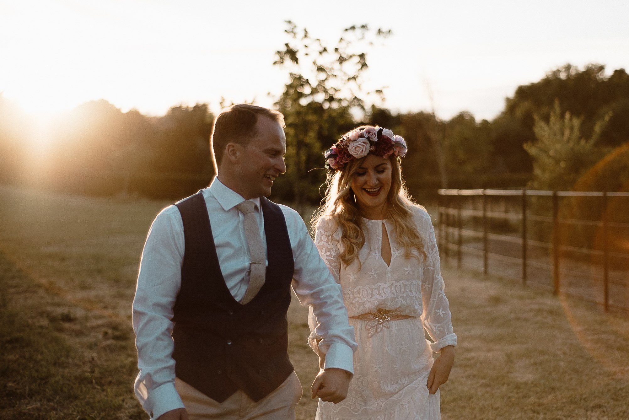 A smiling couple, with the man in a white shirt and vest and the woman in a white lace dress with a flower crown, walking arm in arm outdoors during sunset.