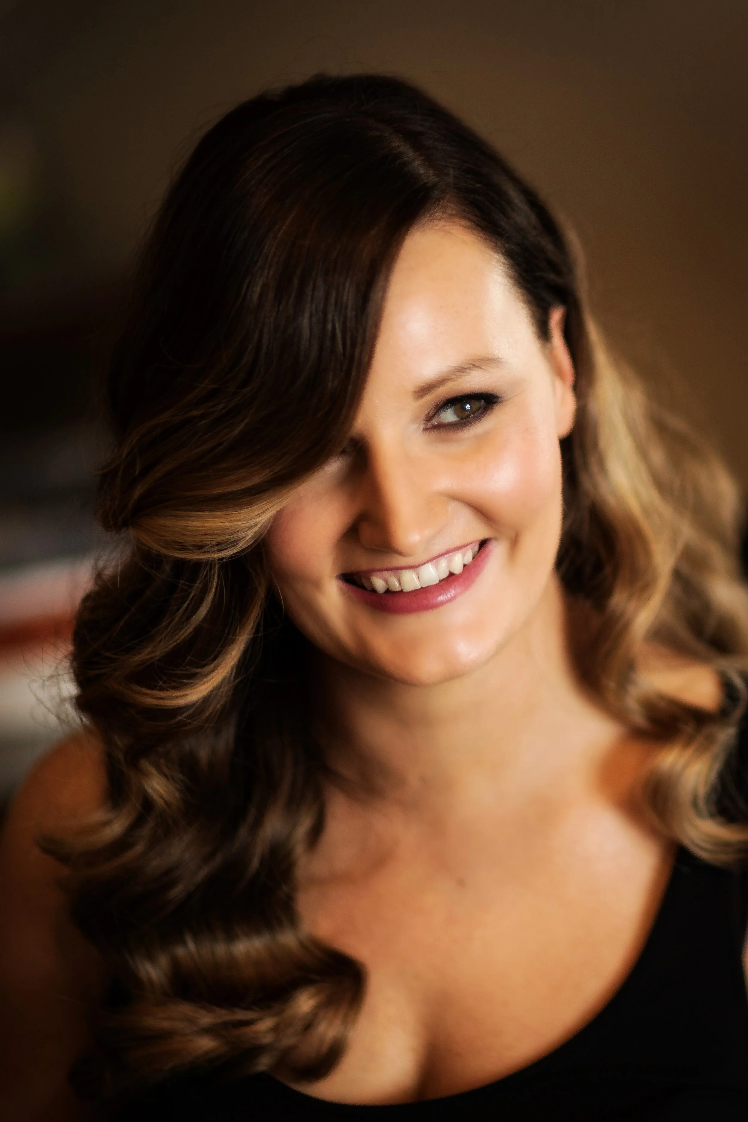 A woman with wavy brown hair and highlights, smiling and wearing a black sleeveless top, indoors with warm lighting.