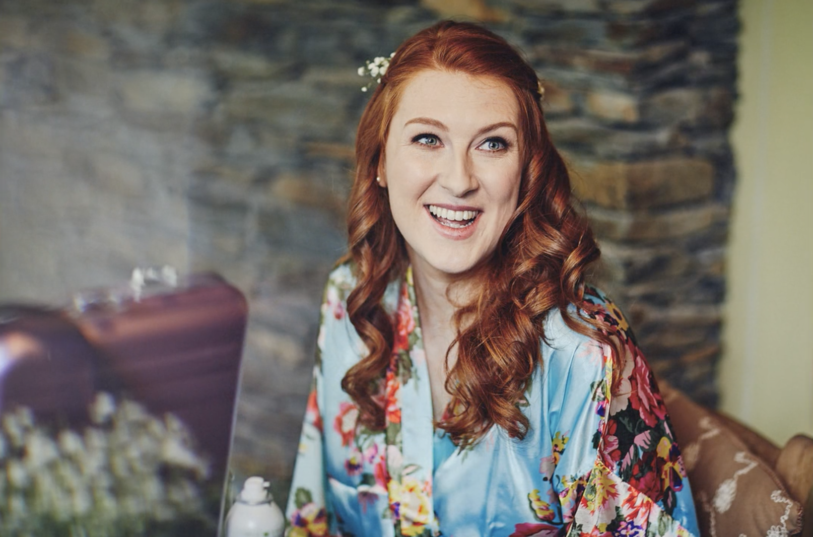 A cheerful woman with curly red hair and a floral robe, sitting indoors near a stone wall, smiling at the camera.