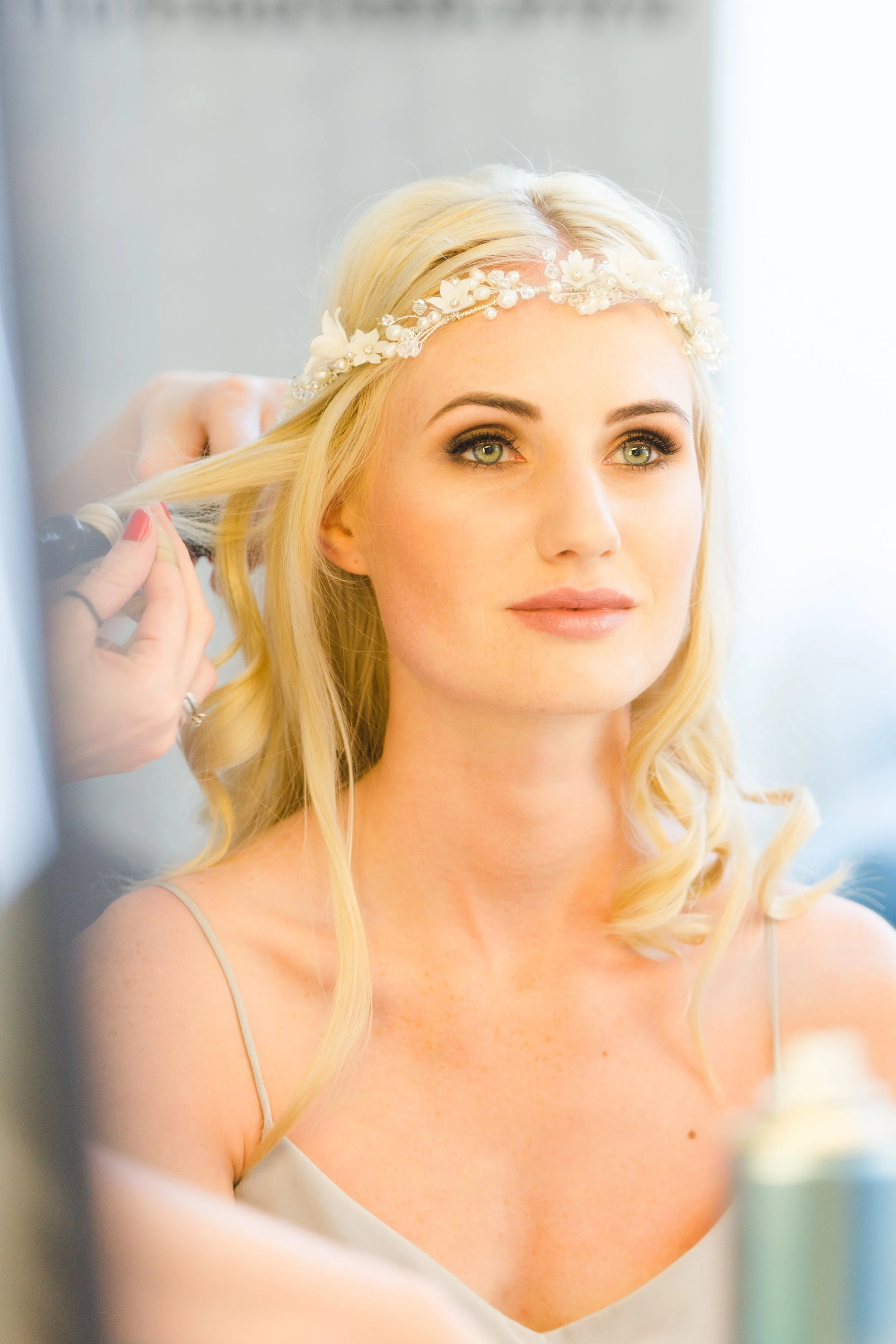 A woman with blonde hair and green eyes wearing a floral headband has her hair styled by a hairstylist.