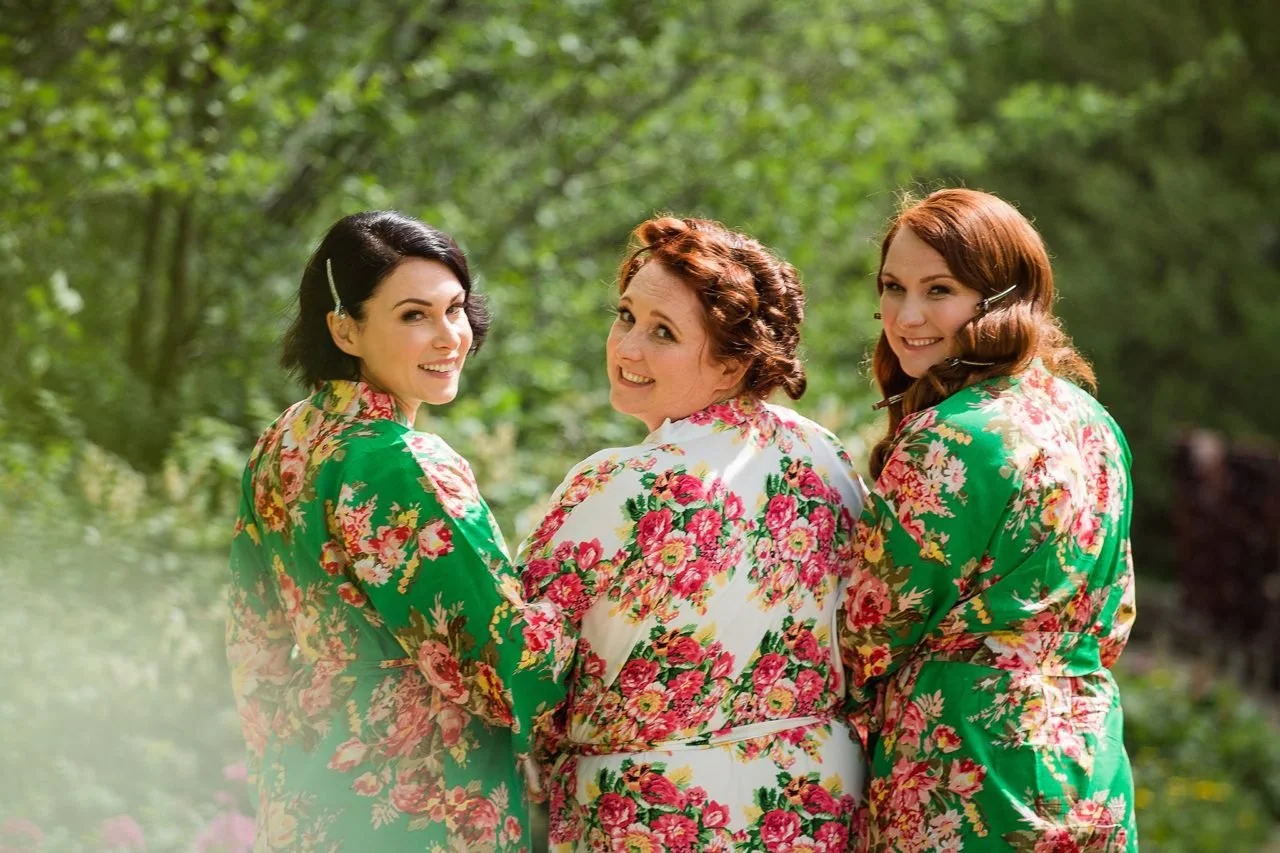 Three women in floral robes smiling and walking through a green, wooded area.