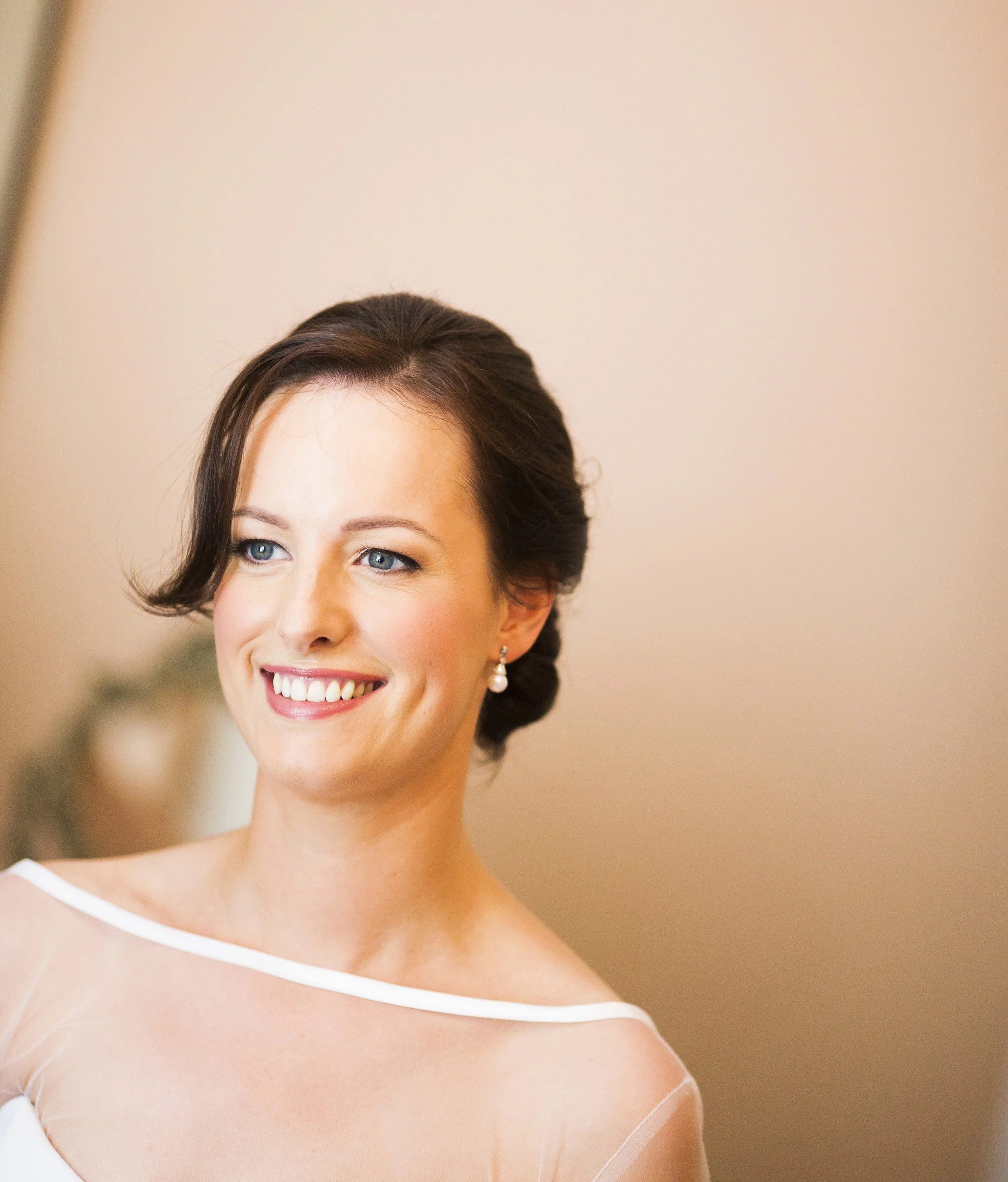 A woman with short brown hair, blue eyes, and pearl earrings smiling softly, wearing a white dress with a sheer overlay.