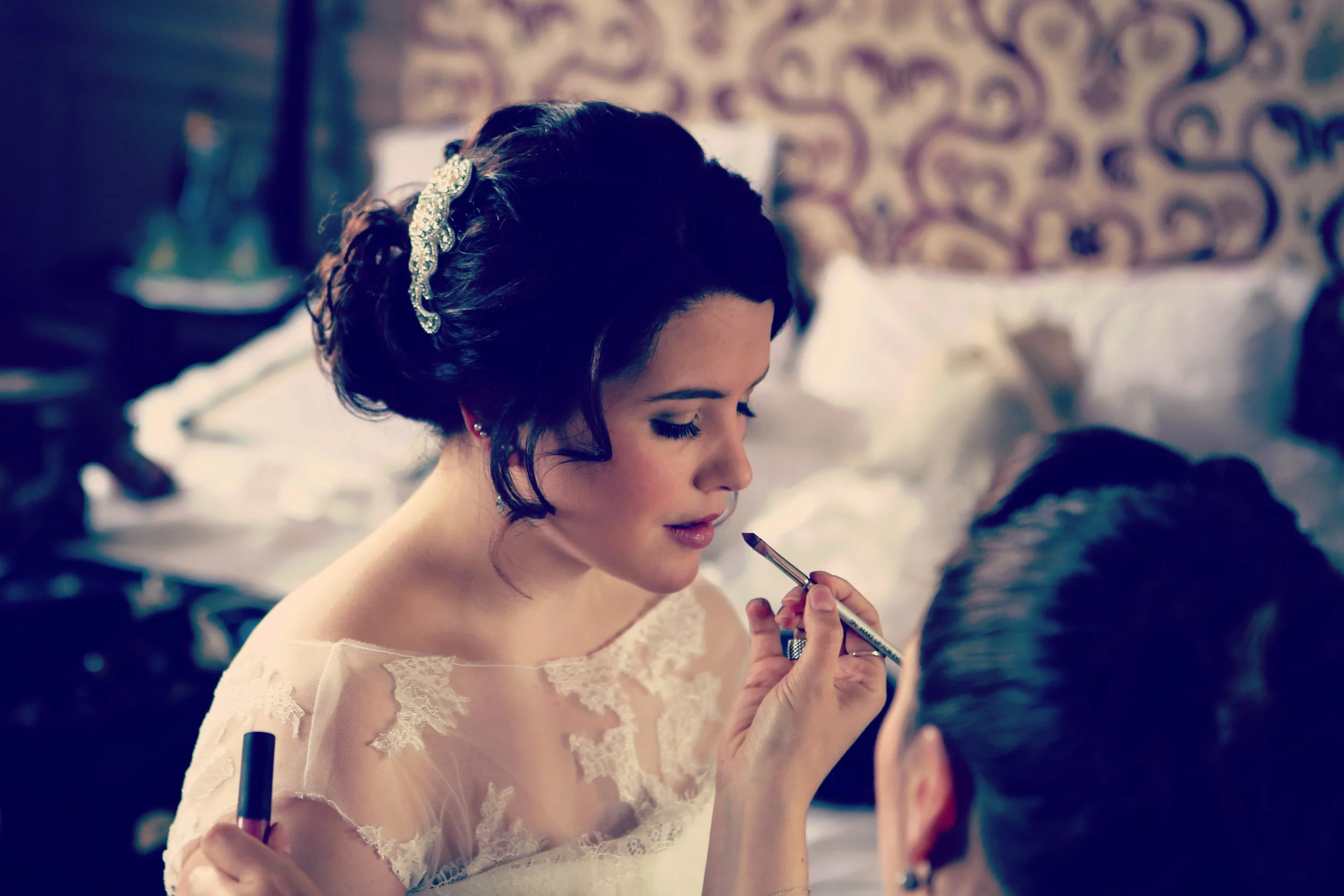Bride getting her makeup done, in a lace dress, with a decorative hairpiece, sitting in a room with patterned wallpaper.