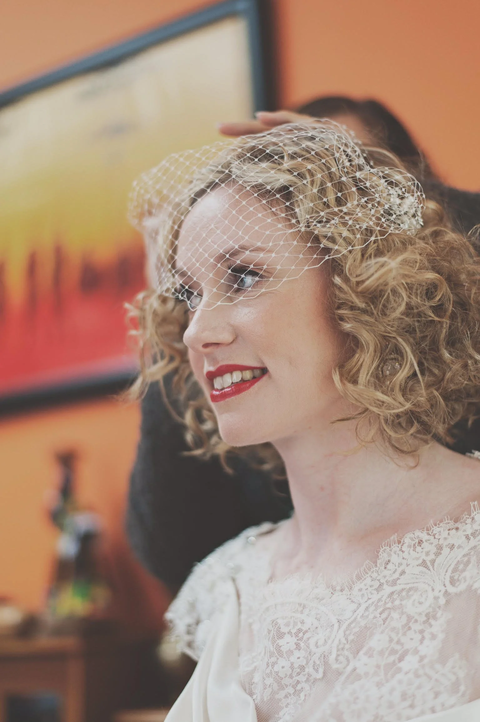 A woman with curly blonde hair, wearing a white lace top, is smiling as she gets ready for her wedding, with a veiled headpiece.