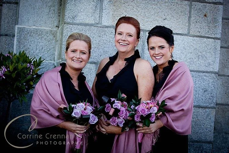 Three women smiling outdoors, each holding bouquets of purple and pink roses, dressed in black and pink shawls.