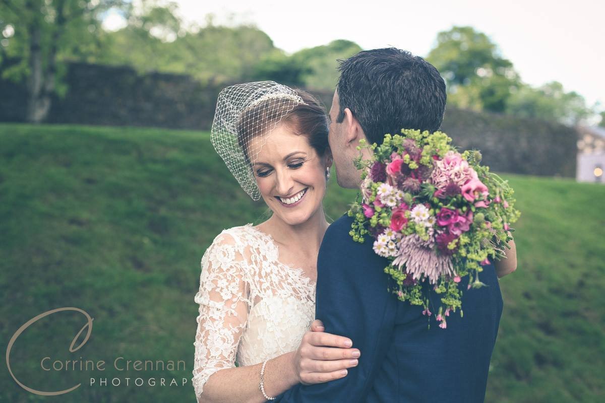 A bride and groom share a moment outdoors, with the groom holding a large bouquet of pink and purple flowers on his shoulder. The bride wears a lace wedding dress and a birdcage veil, smiling with her eyes closed.