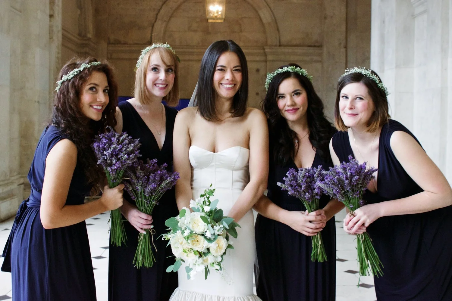 A bride in a white strapless wedding dress holding a bouquet of white roses and greenery, standing with four bridesmaids dressed in navy blue, holding purple lavender bouquets, inside a large stone building with high ceilings and an arched entrance.