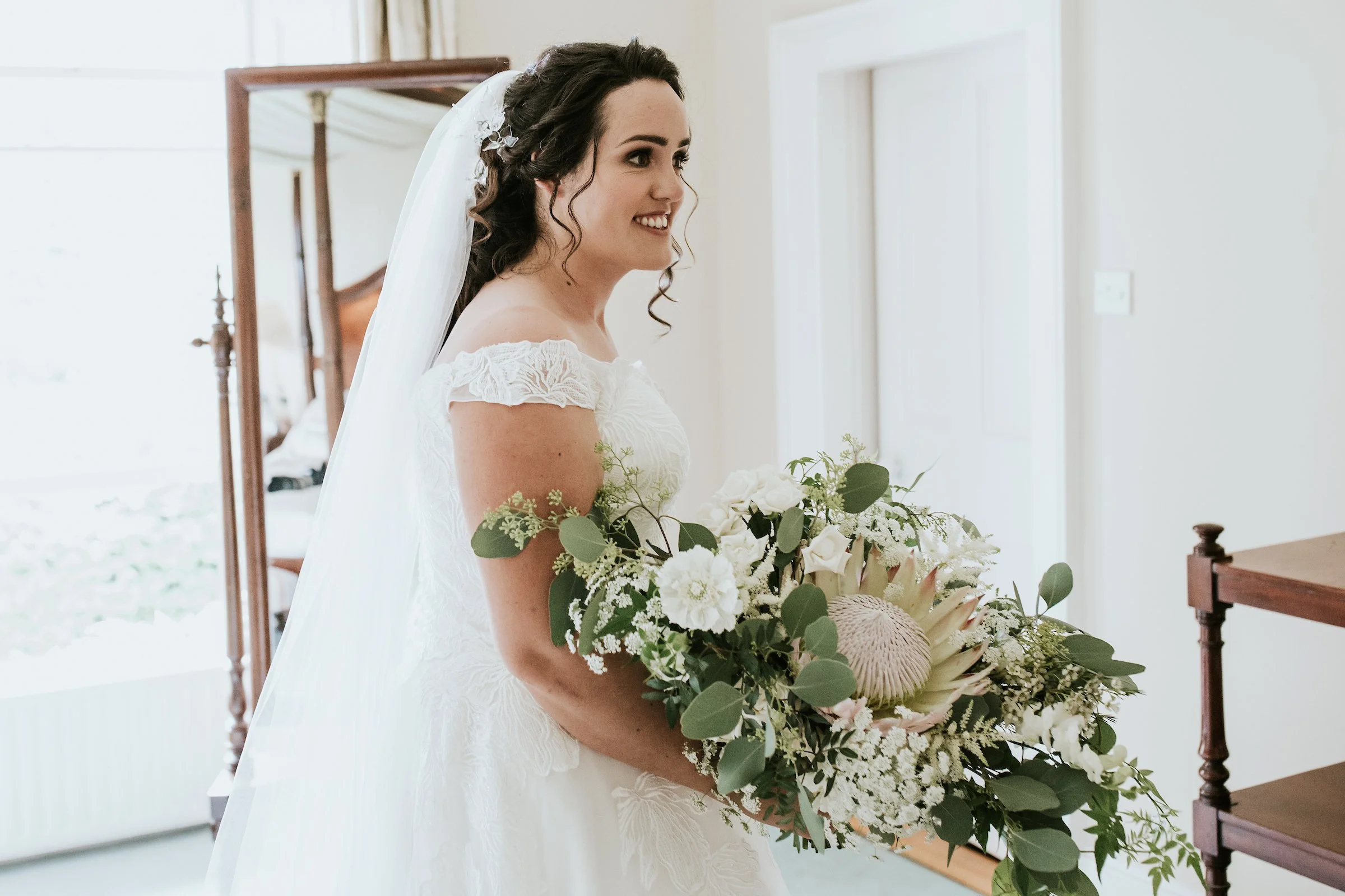 A bride in a white wedding dress holding a large bouquet of white and green flowers, standing indoors with a mirror and wooden furniture in the background.