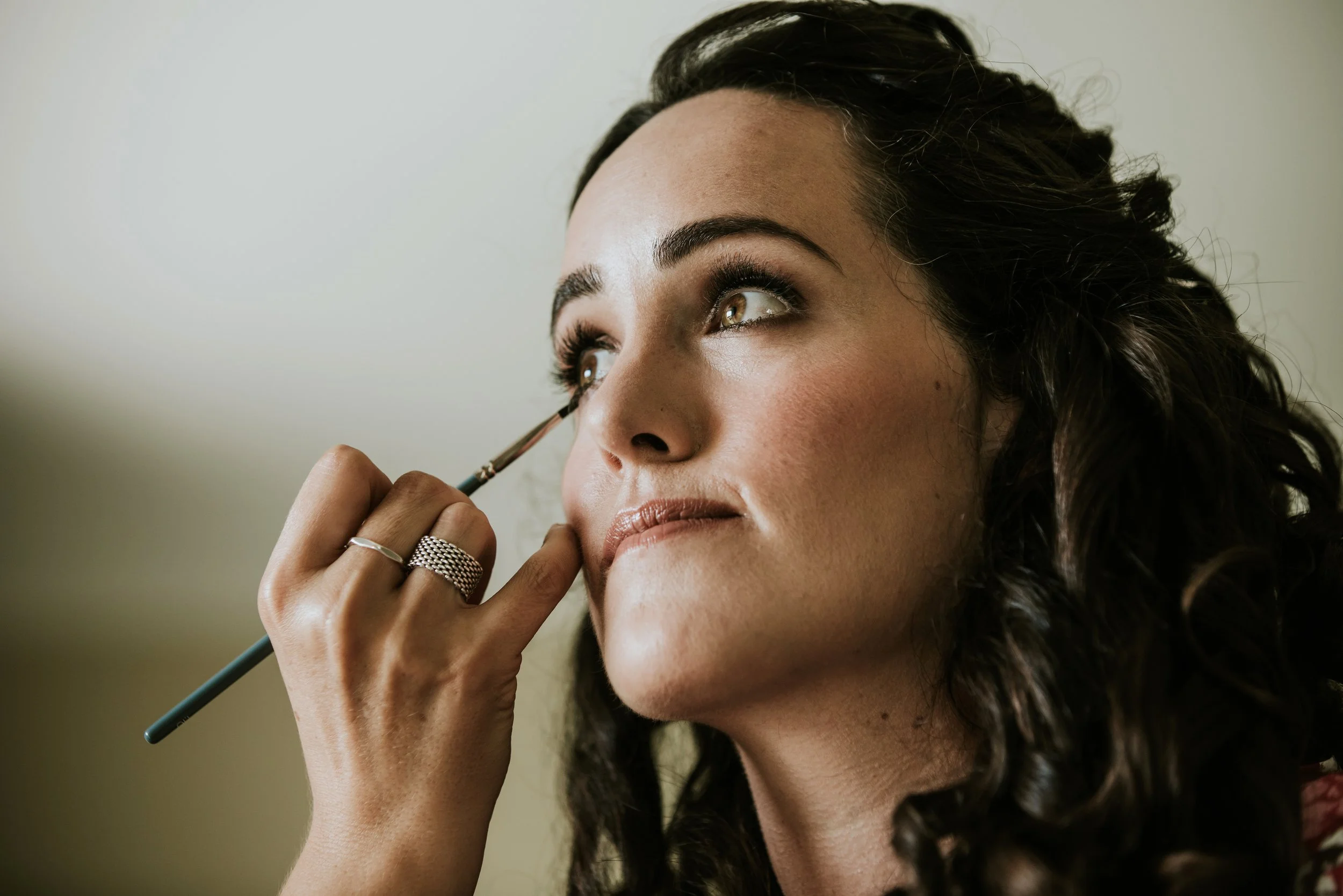 A woman with dark, curly hair and makeup is applying mascara with a brush, looking slightly upward.