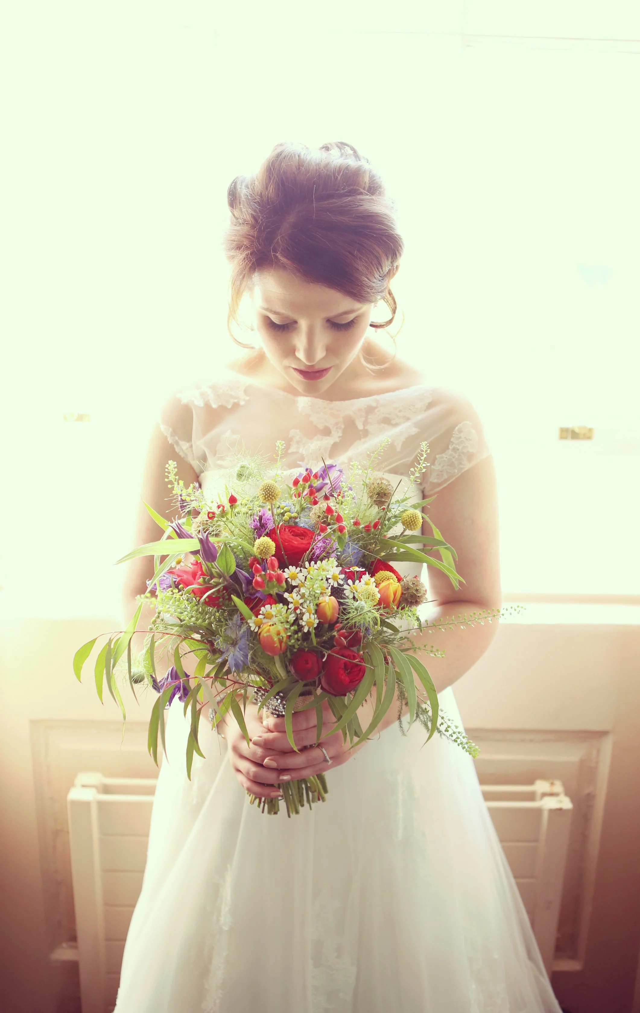 A bride in a white dress holding a colorful bouquet of flowers, standing indoors near a bright window.