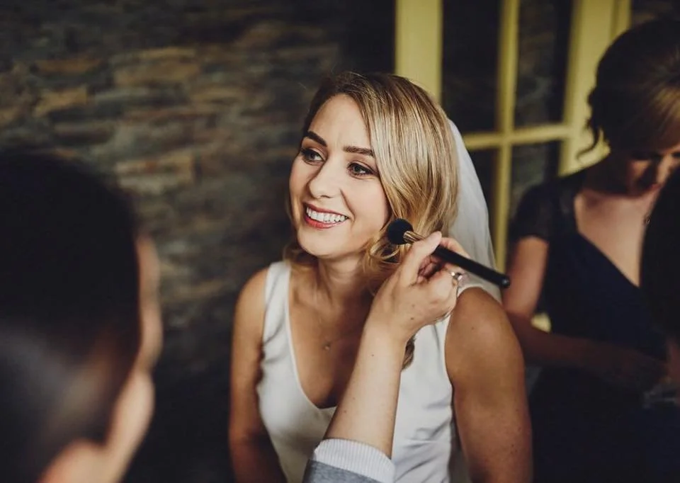 A woman in a white dress smiling as she gets her makeup done with a brush by a makeup artist, with another woman in black behind her and a stone wall in the background.
