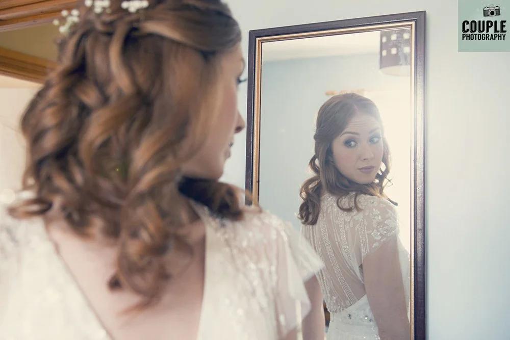 A woman with styled brown hair is looking at her reflection in a mirror, with a soft expression. She is wearing a white dress with lace details. The background features a framed mirror and a wall with sunlight streaming in.