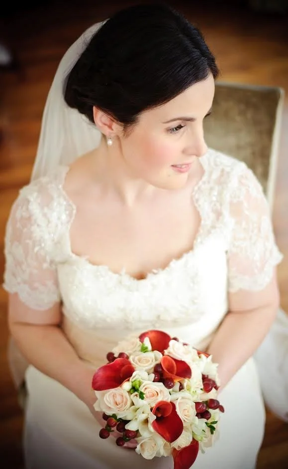 A bride with dark hair in a wedding dress and veil, holding a bouquet of red and white flowers, sitting on a chair indoors.
