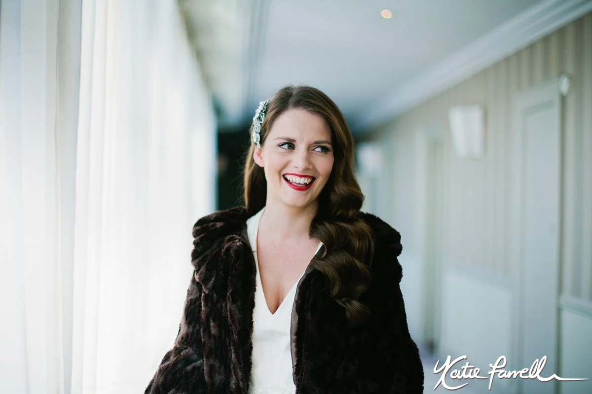 A woman with long curled hair, wearing a white top and a dark fur coat, smiling and standing in a hallway with light-colored walls and curtains, with a signature at the bottom right reading "Yette Farrell."