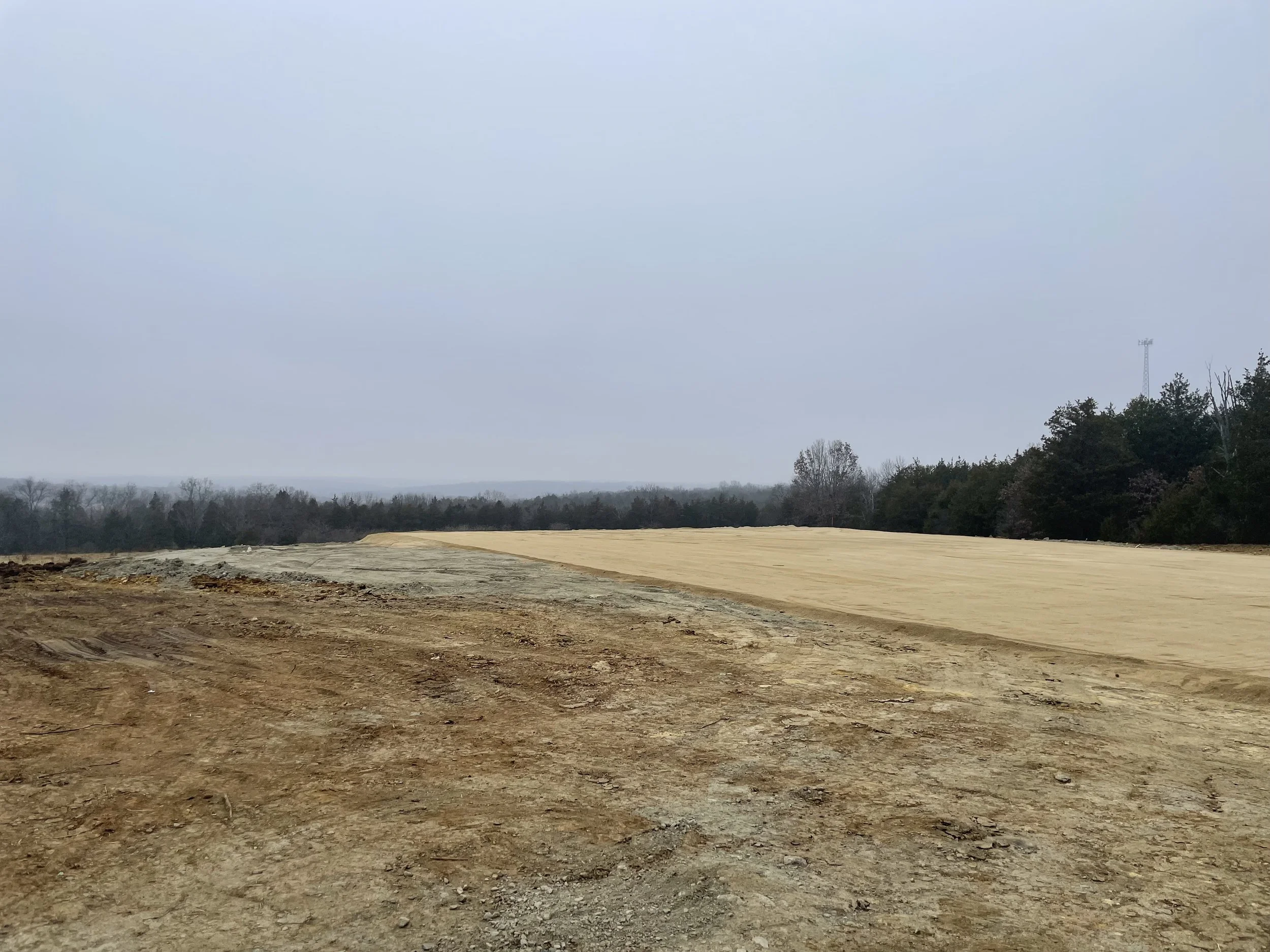A construction site with freshly leveled dirt and gravel, with trees and an overcast sky in the background.