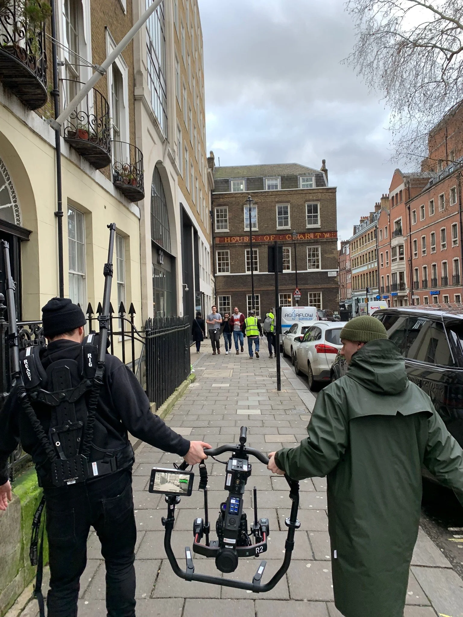 Two men, one in black with camera equipment and the other in green raincoat, hold a camera crane on a city sidewalk with parked cars, buildings, and pedestrians in the background.