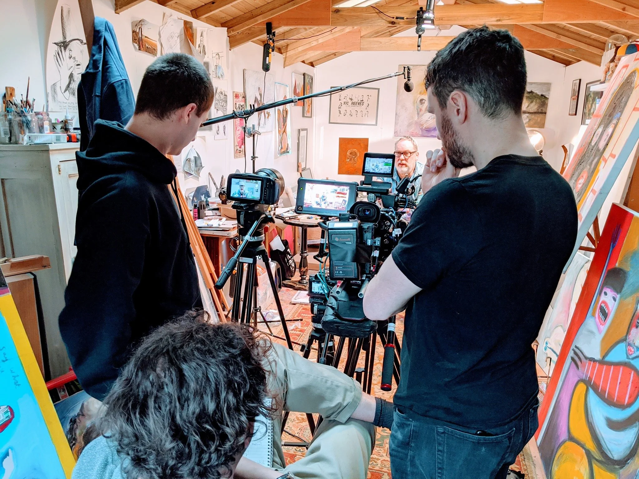 A film crew is filming inside an art studio, with artists working on paintings and sketches on the walls and easels. Crew members operate cameras and equipment while a person appears to be getting ready for a shot.