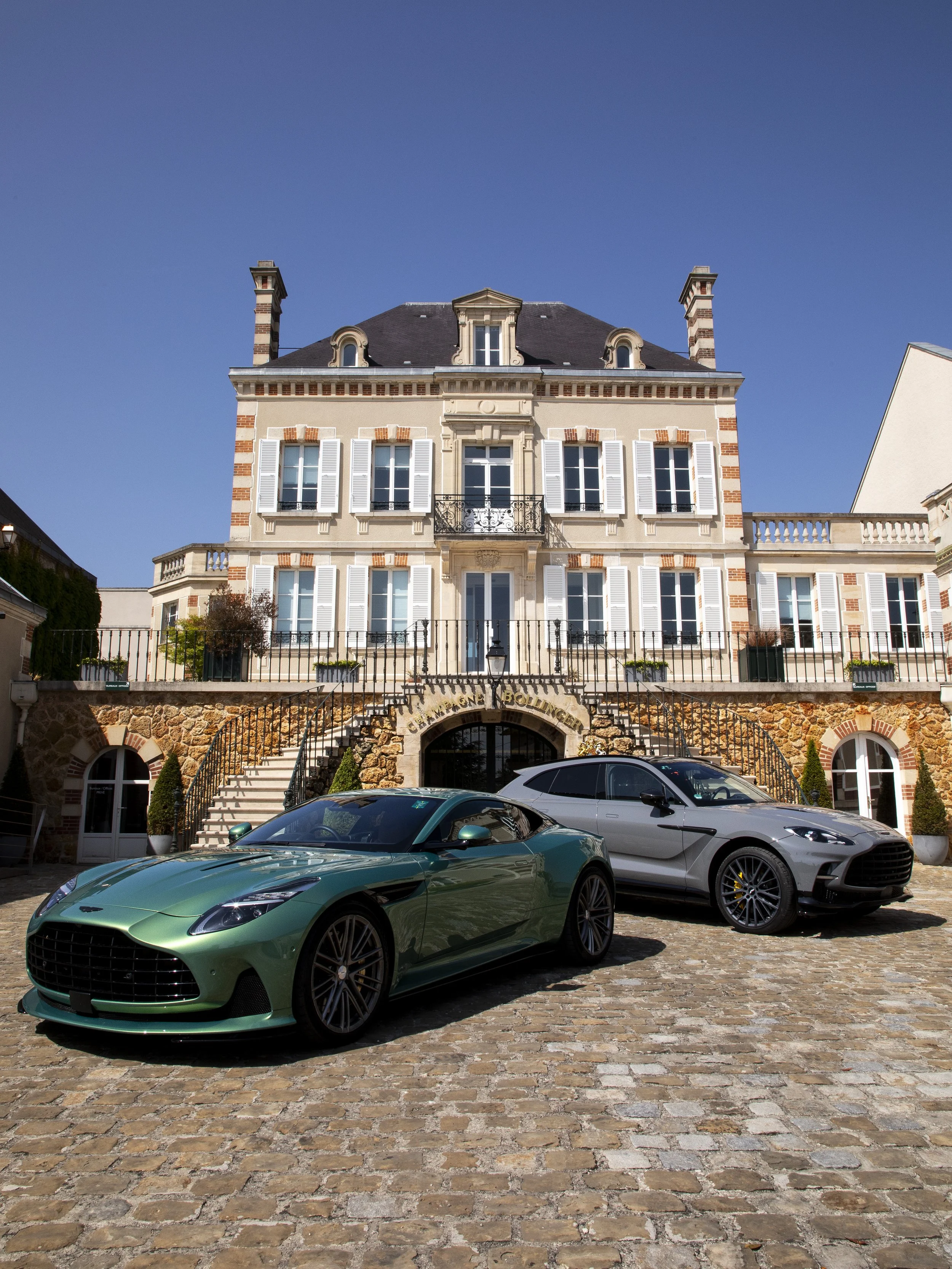 Luxury villa with stone and stucco architecture, two luxury cars parked on cobblestone driveway, clear blue sky.