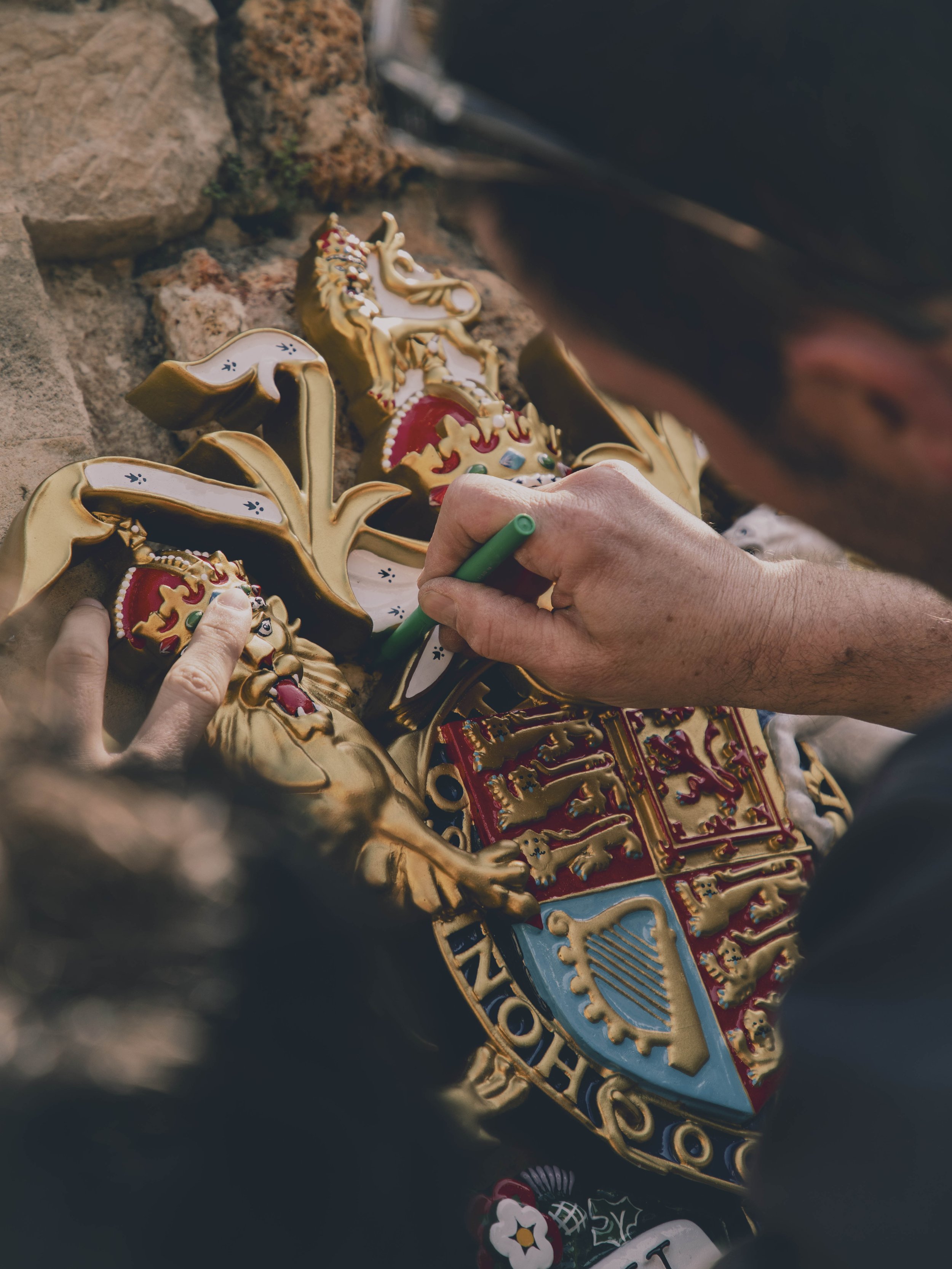 A person is painting gold and colorful decorative ceramic or plaster plaques with heraldic emblems and lions, mounted on a stone wall.
