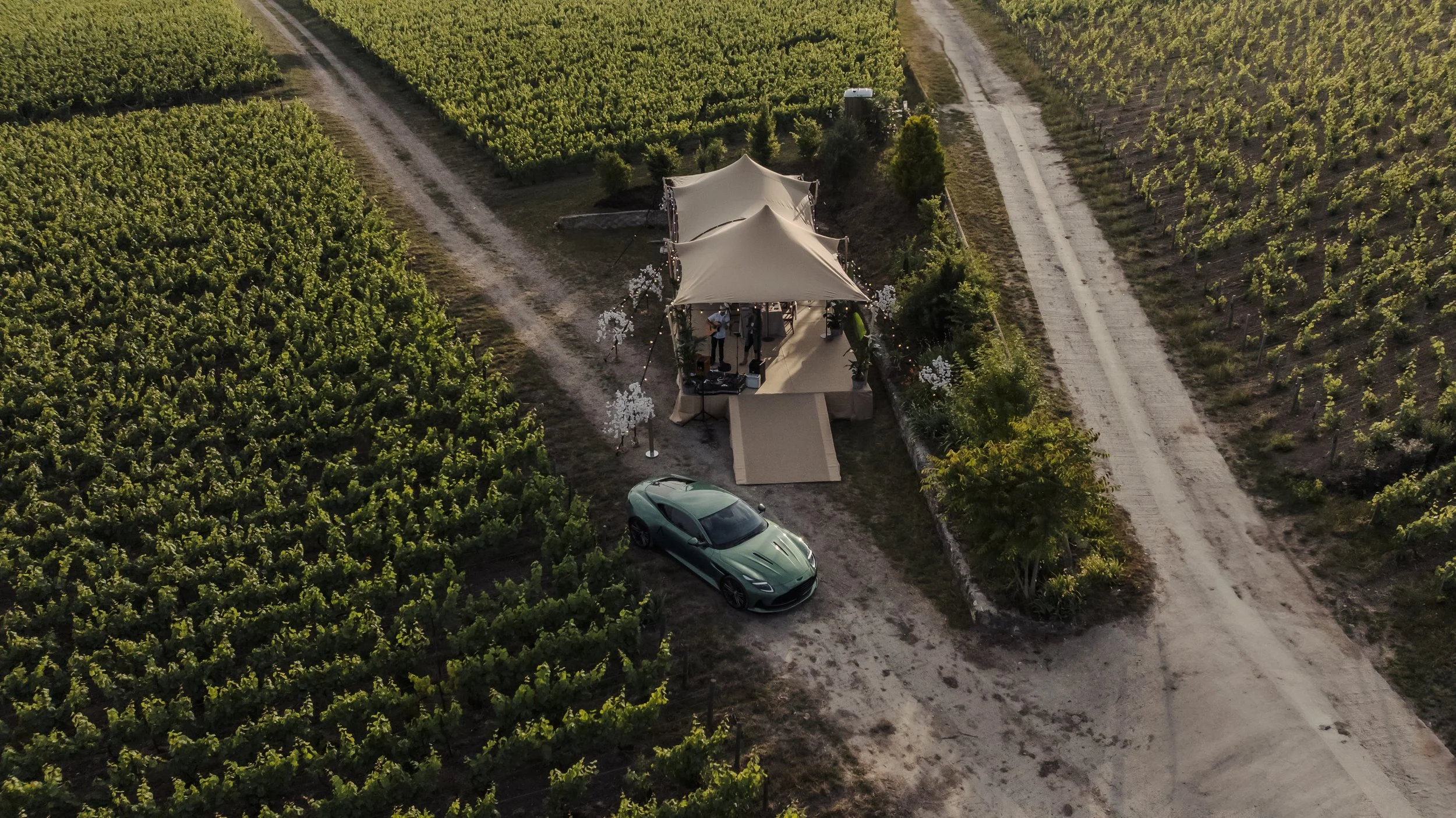 A drone shot of a wedding ceremony setup in the middle of a vineyard, with a small stage, two canopy tents, and a green sports car parked nearby.