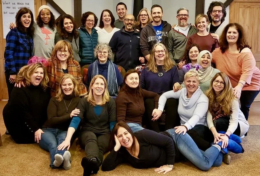 Group of approximately 22 diverse smiling people gathered closely together in an indoor setting, appearing joyful and friendly.