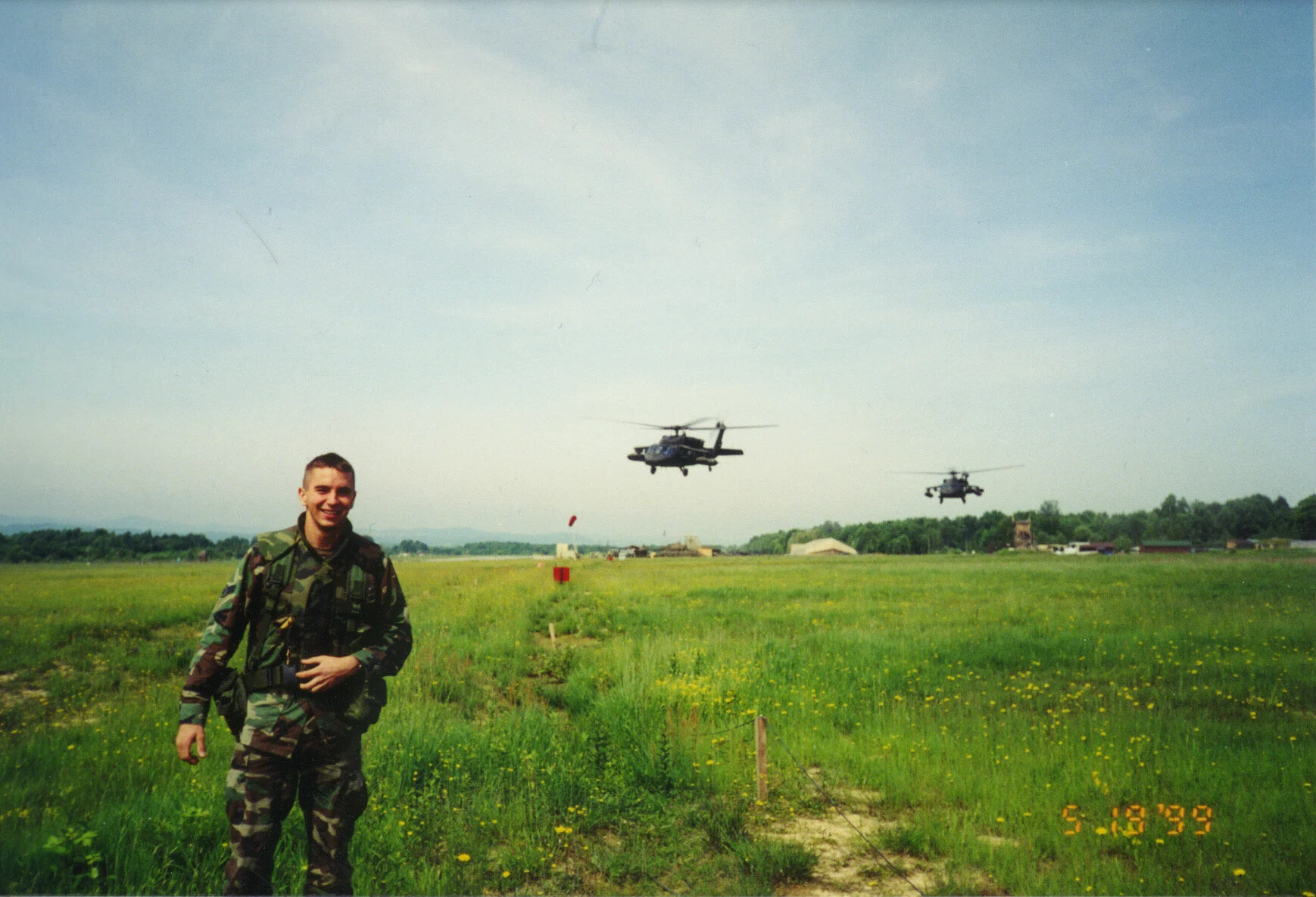 Conrad Hayter, leadership coach and facilitator, in military uniform standing in a grassy field with yellow flowers, with two helicopters flying overhead in the background, and small buildings and trees in the distance.