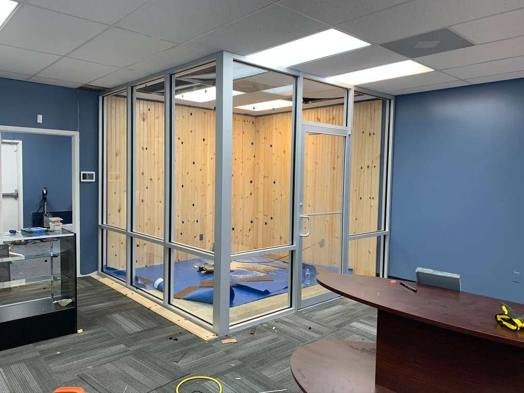 Room under construction with wooden walls, glass framing installed by Glassworks, and tools on scattered on the dark gray carpeted floor.