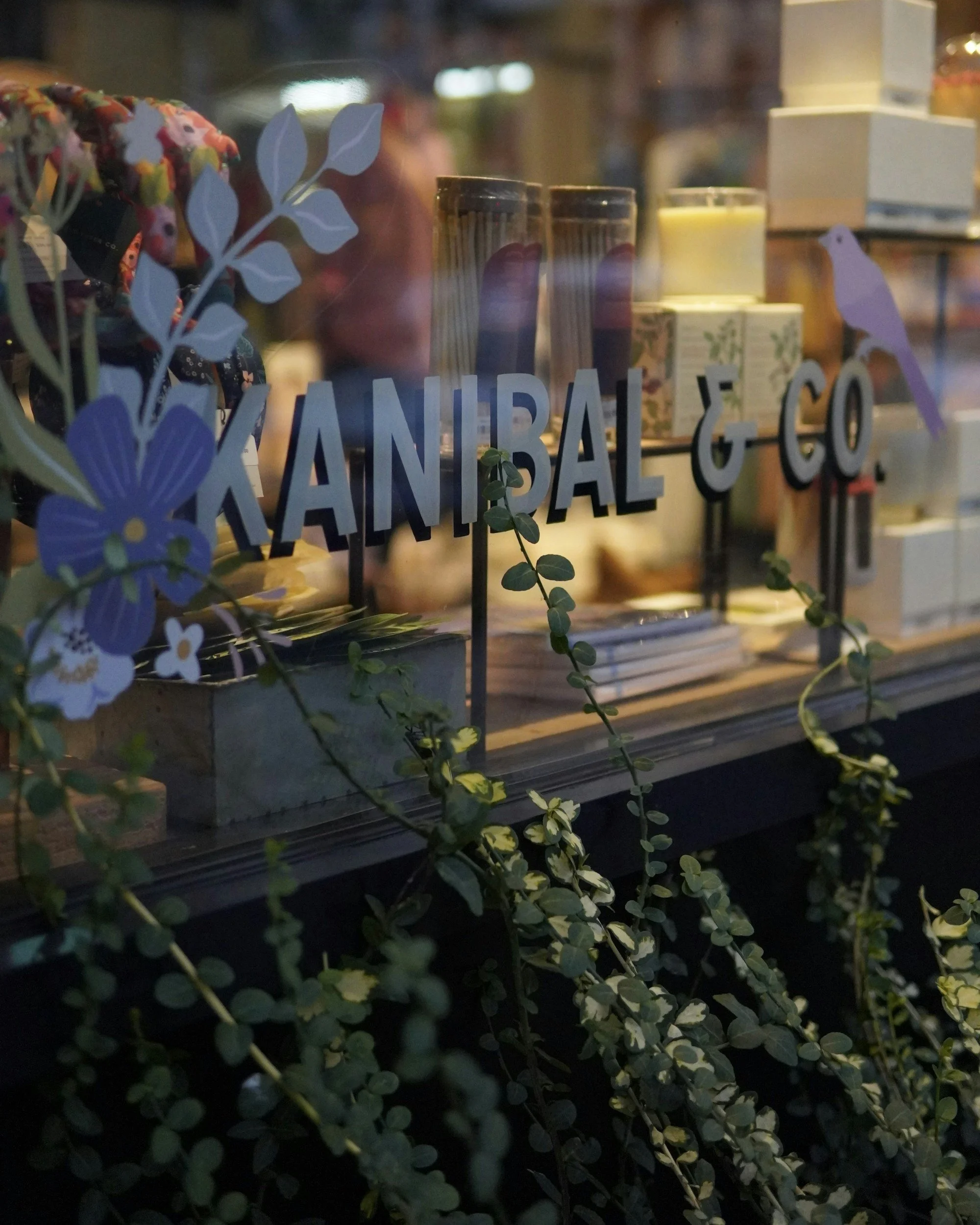 Store window with the sign 'KAINBAL & CO.' installed by Glassworks surrounded by decorative plants and flowers, displaying candles and boxes inside.