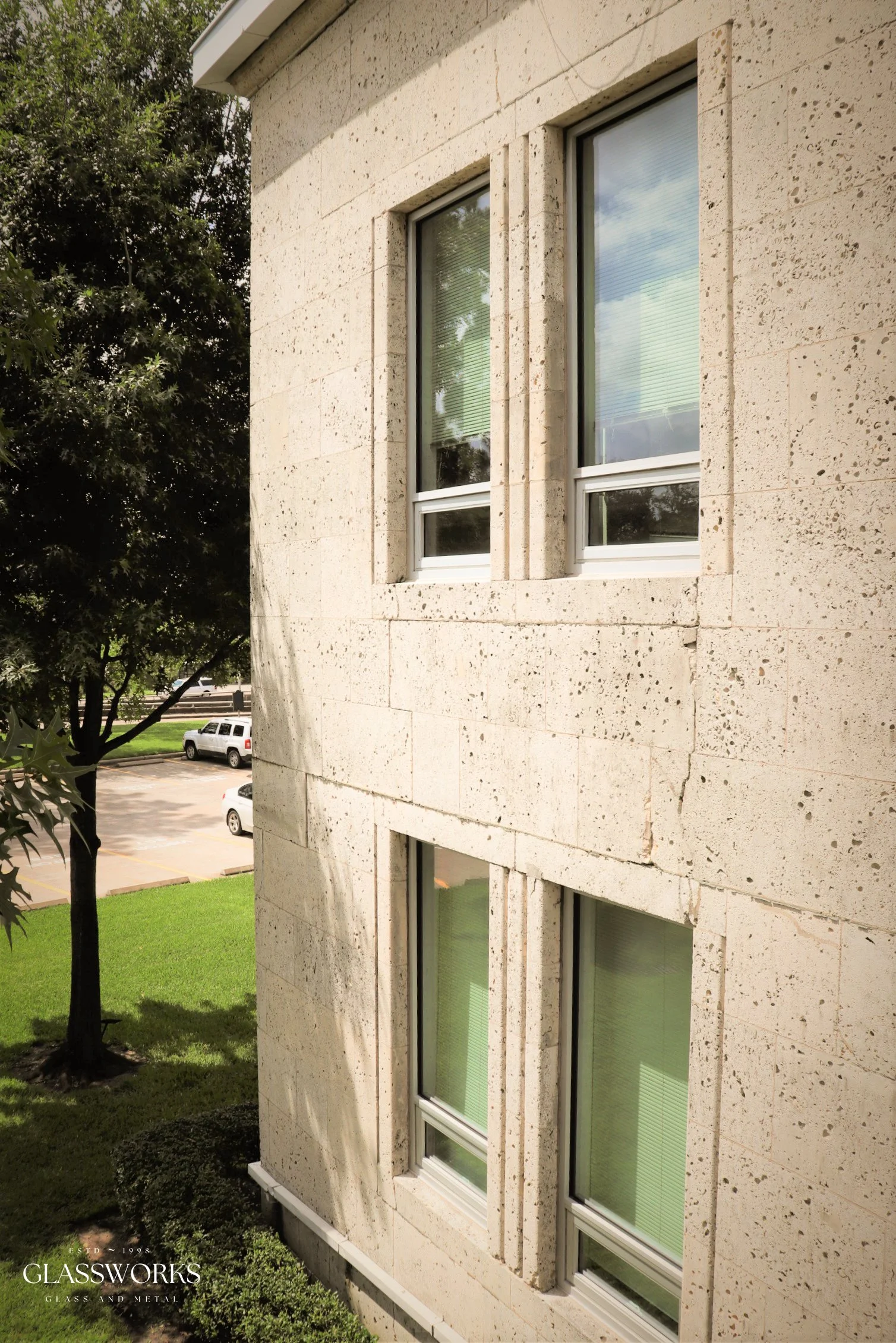 Exterior view of a beige stone building with four rectangular windows, two on each floor, with white frames and blinds. There's a tree and parked cars in the background, and a logo for Glassworks in the bottom left corner.