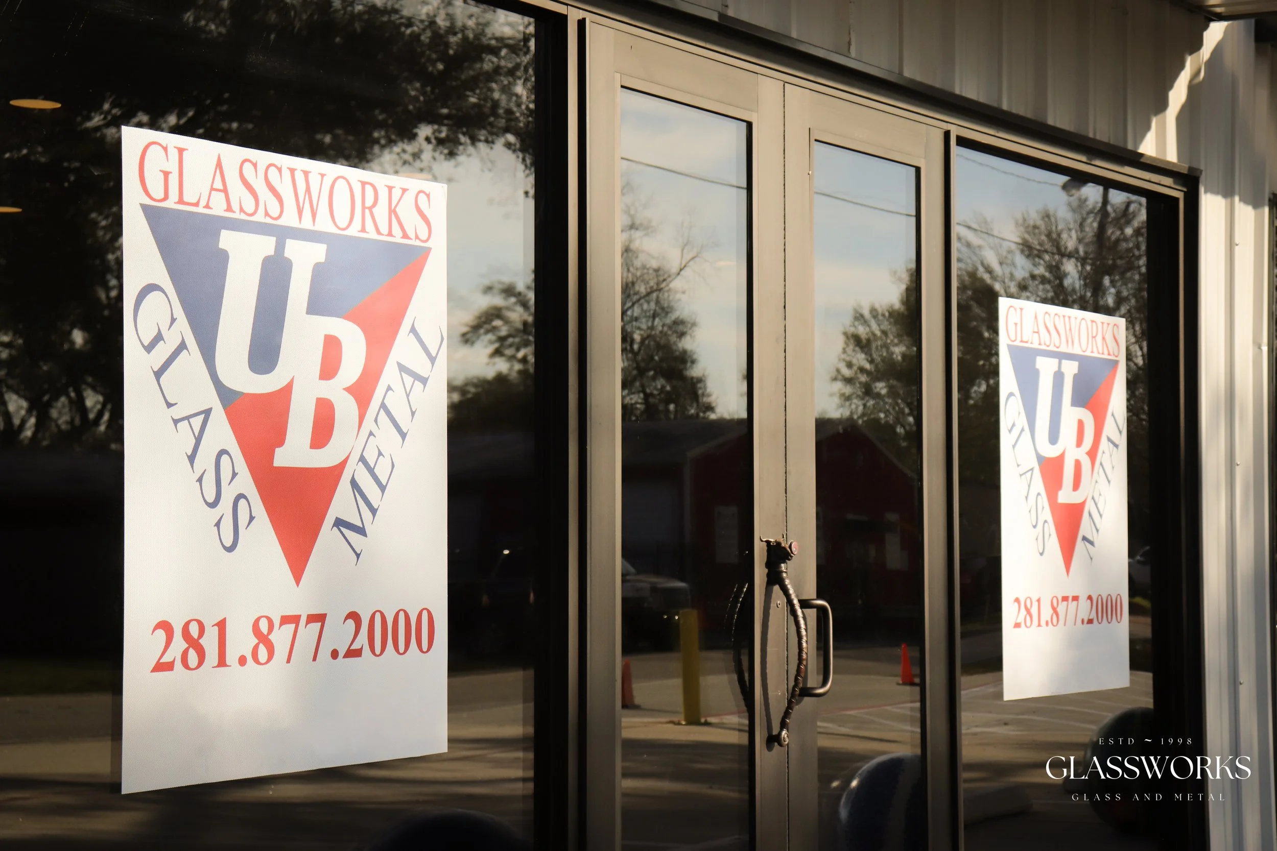 Glassworks storefront window with two signs featuring the company's logo, a red, white, and blue triangle with the initials "GUB" and the text "Glassworks Glass & Metal" along with the phone number 281.877.2000, reflected in the glass door surrounded by trees and a cloudy sky.