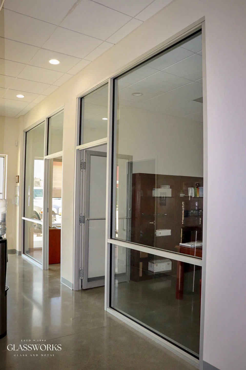 Interior view of an office space with glass partition walls and doors, ceiling lights, and a water cooler on the left.