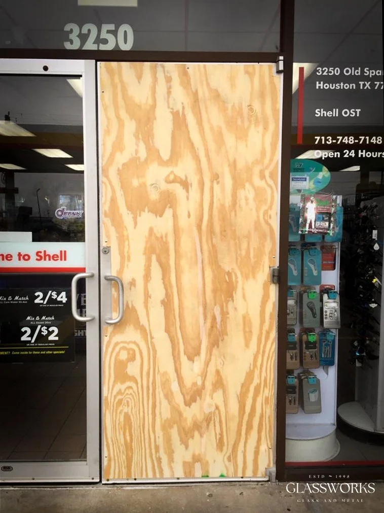A storefront entrance with a plywood door, metal handles, and nearby signs advertising Shell gas prices, a phone number, and a 24-hour service. The glass storefront reflects the interior of the store, including fuel prices and promotional signs.