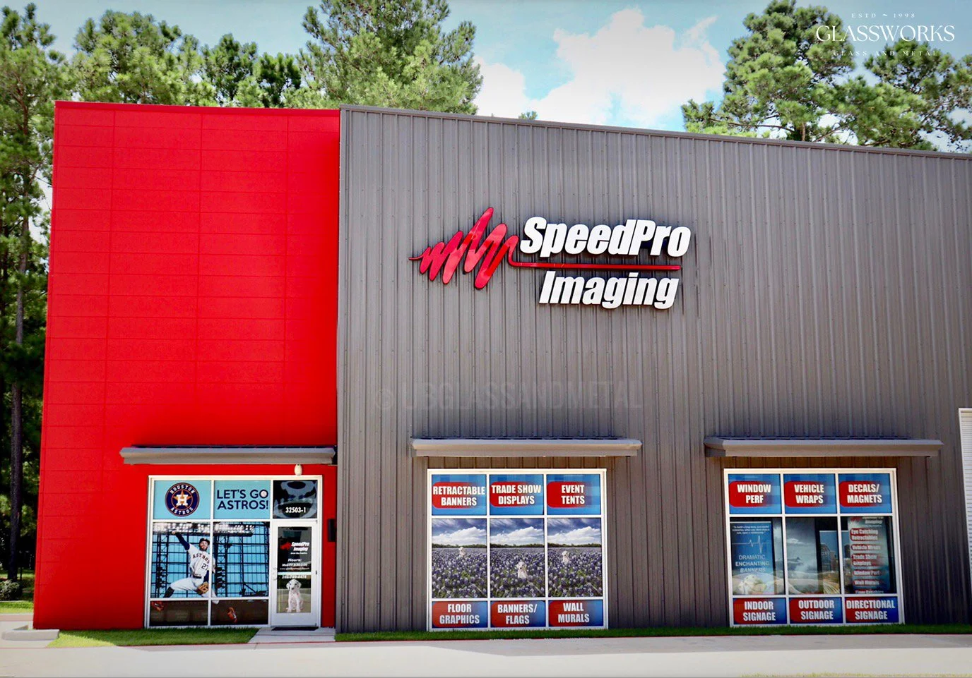 Exterior of a building with the sign 'SpeedPro Imaging' on the front, featuring red and gray panels. The building has windows displaying advertisements for retractable banners, trade show displays, event tents, window performance, vehicle wraps, decals/magnets, indoor and outdoor signage, and directional signage. Trees and a blue sky with clouds are in the background.