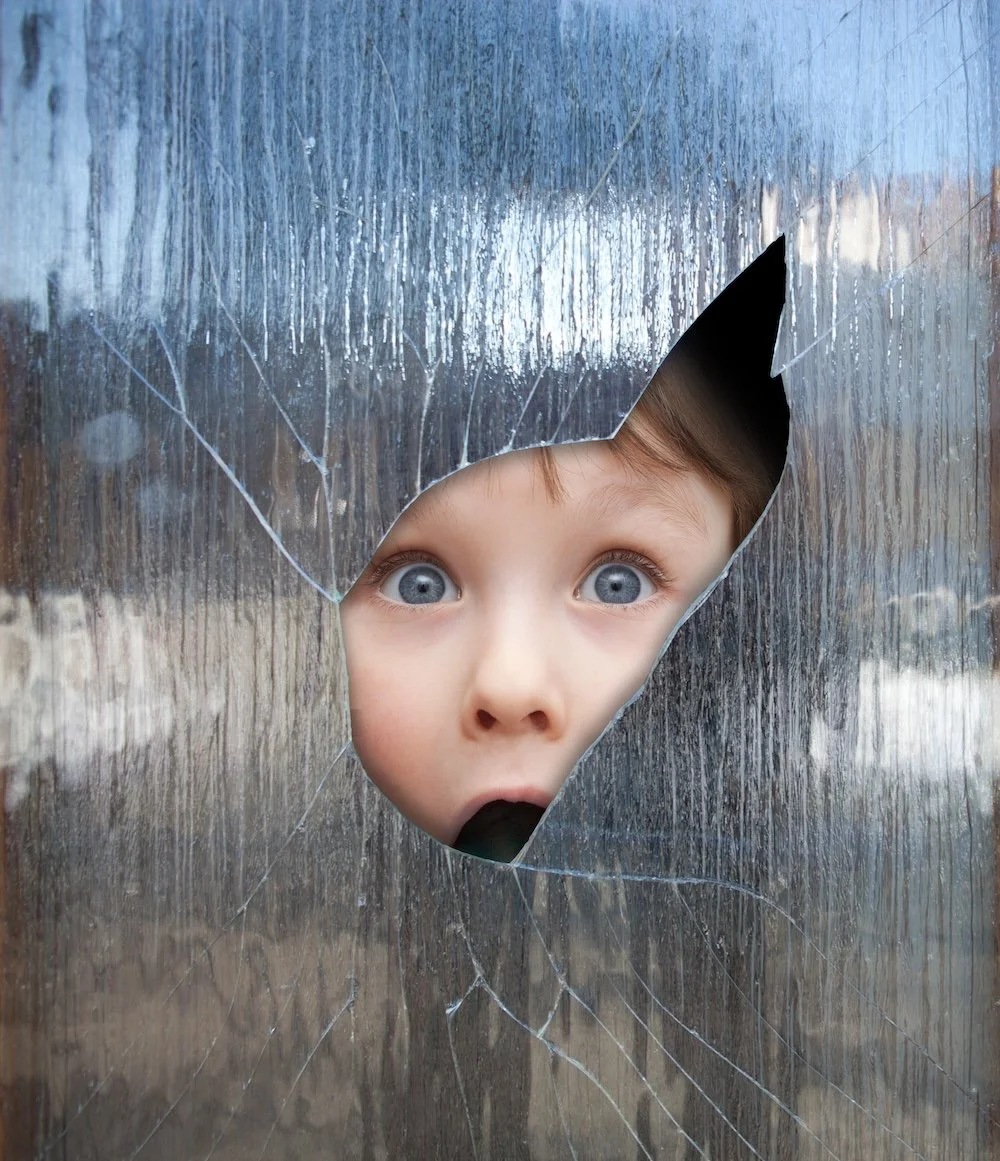 A young child with blue eyes looking through a jagged hole in a frosted glass window.