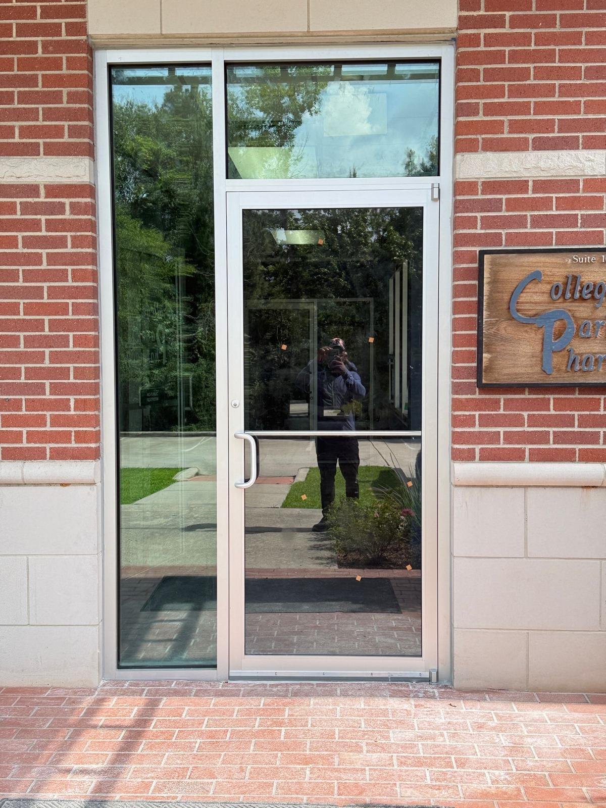 Glass door with metal frame leading into a building, reflecting trees, sky, and a person taking a photo.