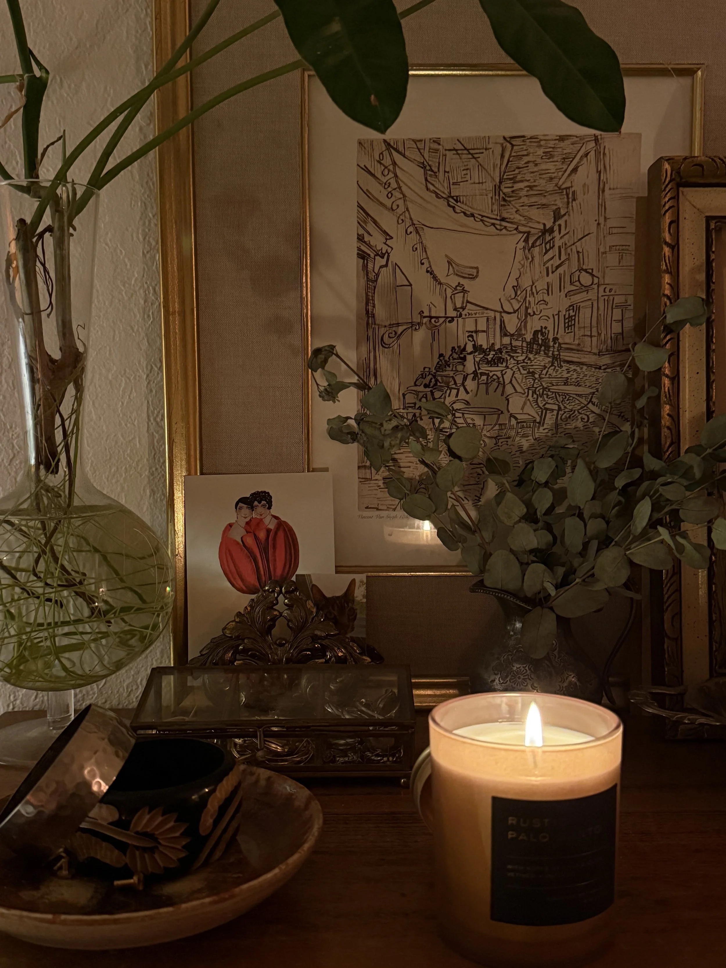A cozy tabletop with a lit candle, decorative objects, framed artwork, and green plants arranged behind them in a warm, softly lit setting.
