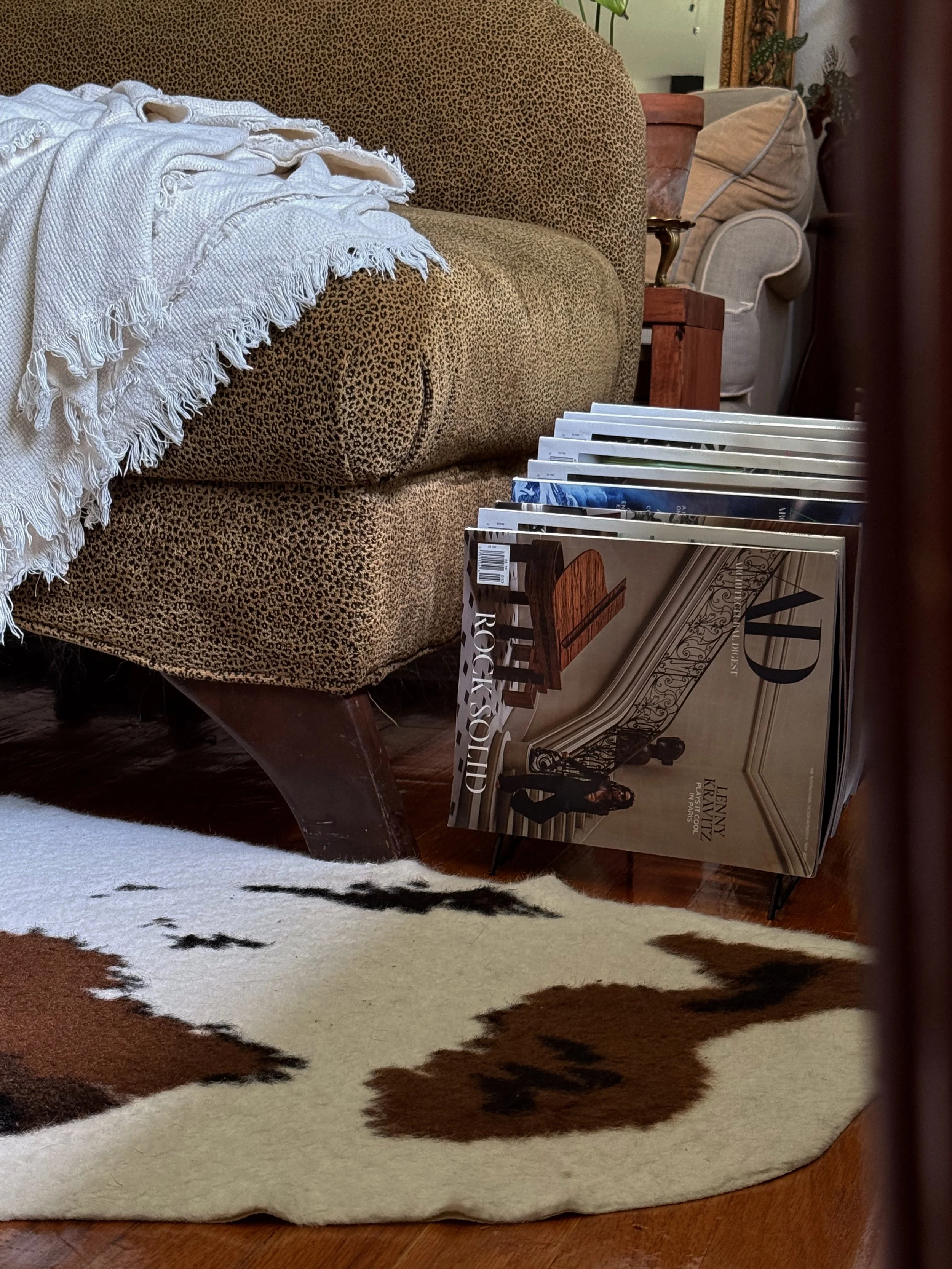 A cozy living room corner with a leopard print couch, a white fringed throw blanket, a magazine stand with magazines, and a cowhide rug on polished wooden flooring.