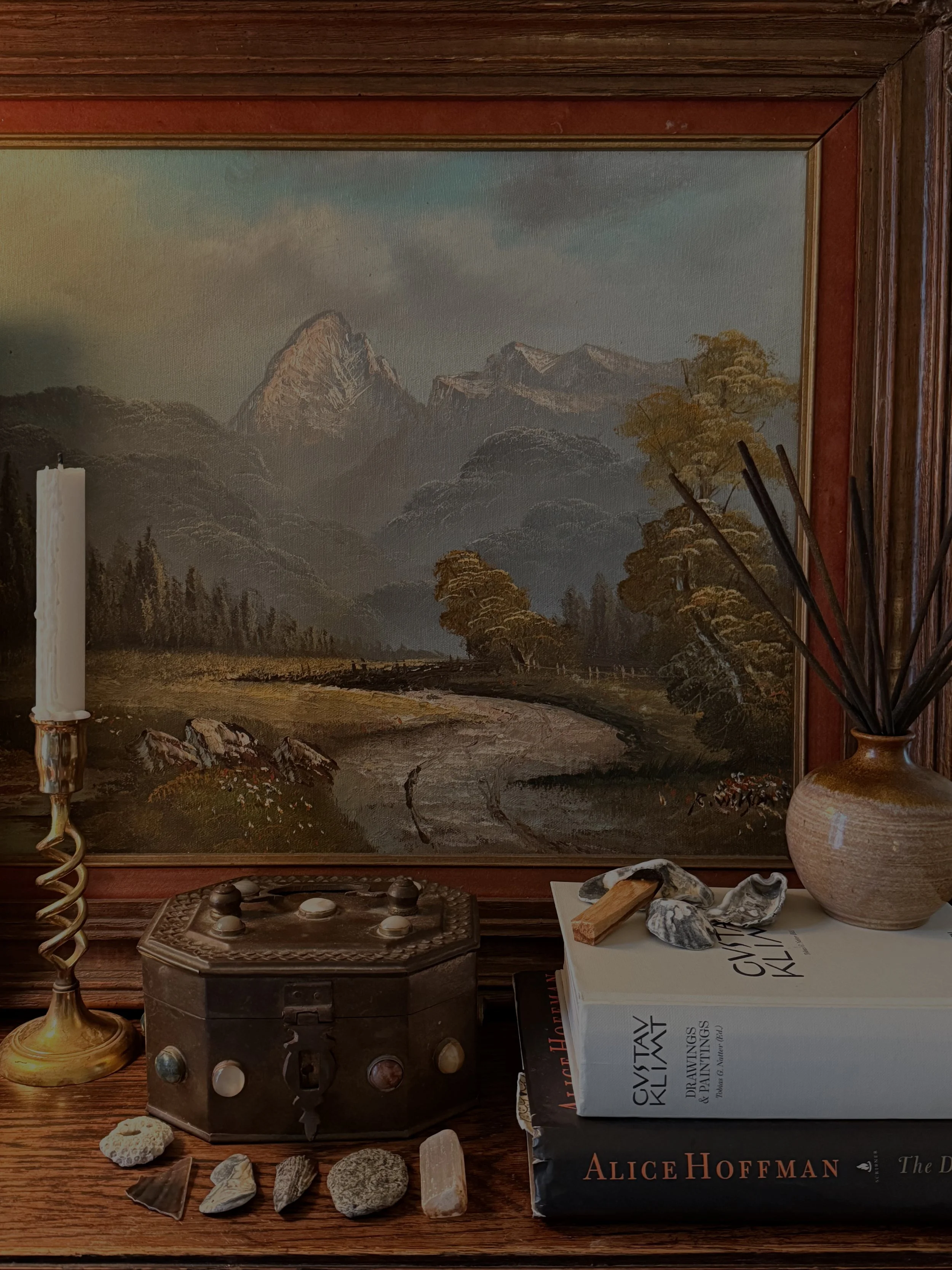 A still life scene with a framed landscape painting of a mountain valley, a candlestick with a white candle, a decorative box, books, a small vase with dried reeds, and various shells and stones on a wooden surface.